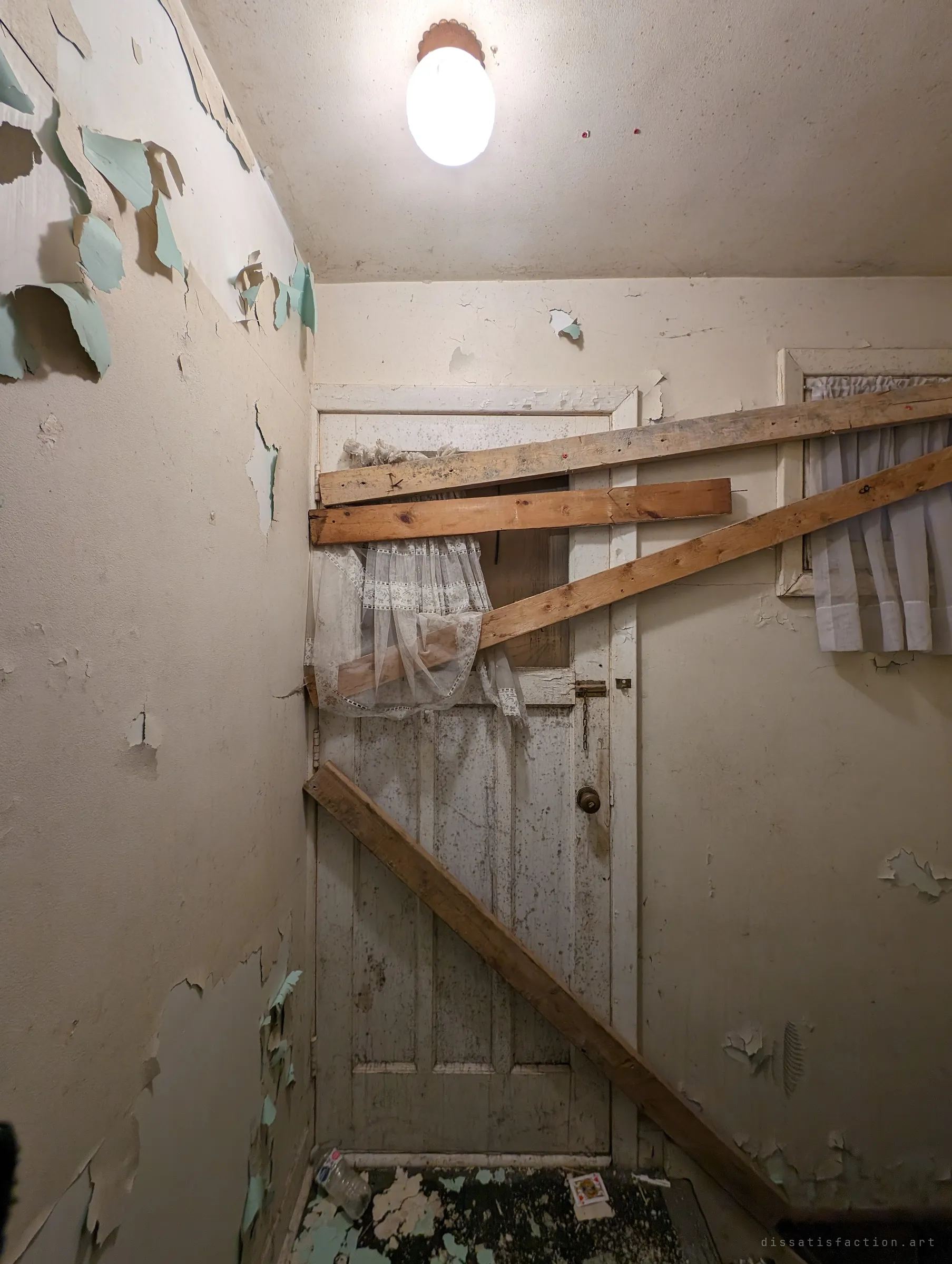A lace-curtained door barricaded with nailed boards in a small hallway, under a glowing ceiling light and walls of peeling mint paint. A playing card lies on the floor.