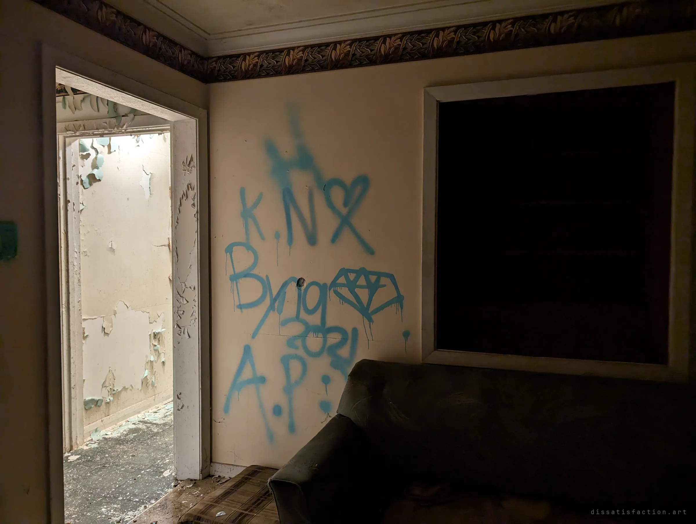 A dim sitting room with blue spray-painted initials, a heart and a diamond on the wall beside a boarded window and a sagging couch. Through the doorway, a hall of peeling paint glows under a single light.