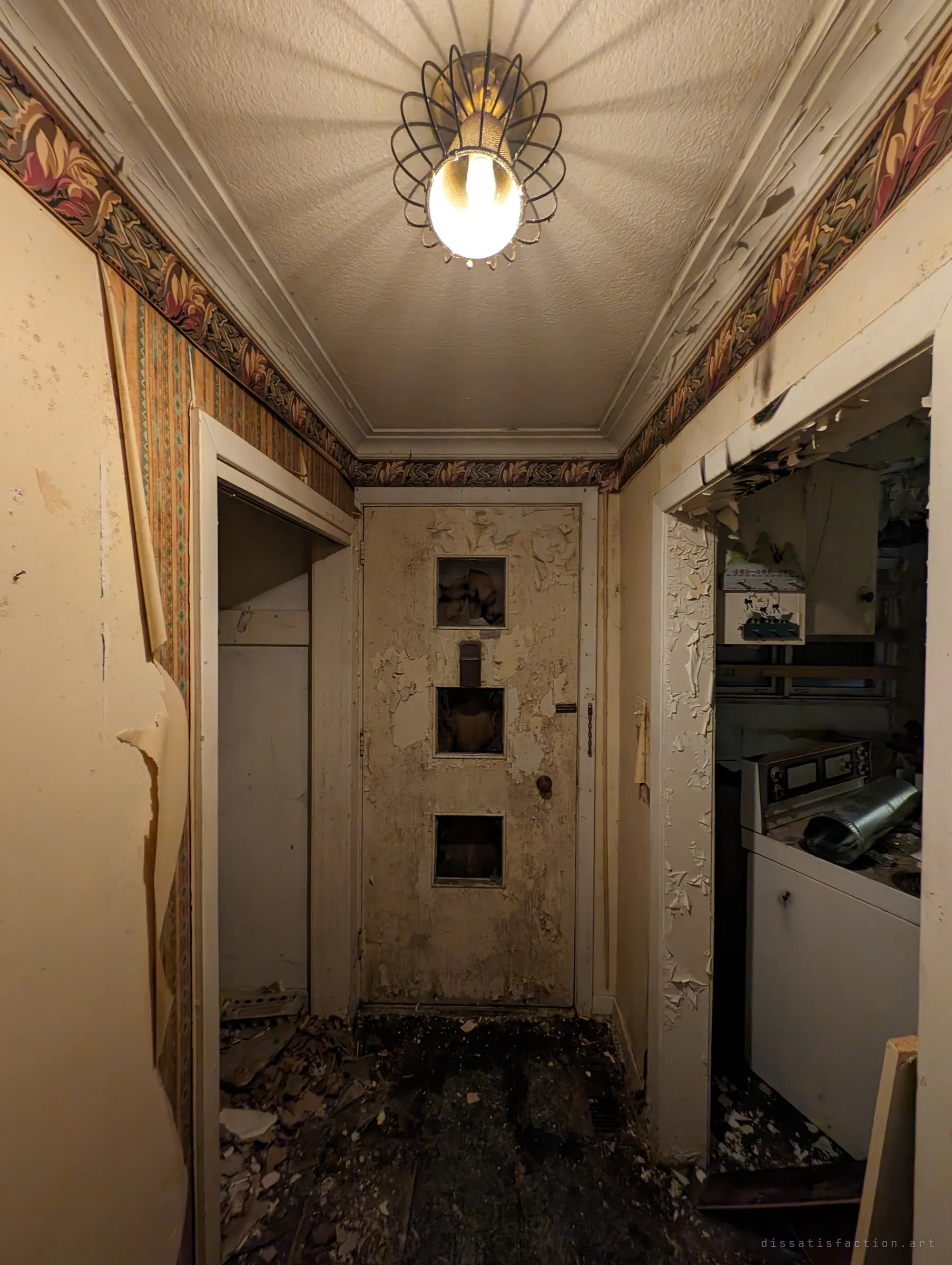A narrow back hall under a caged ceiling light, its patterned border peeling and the paint on the door flaking off in sheets. Through the side opening, a kitchen full of debris and an old stove.