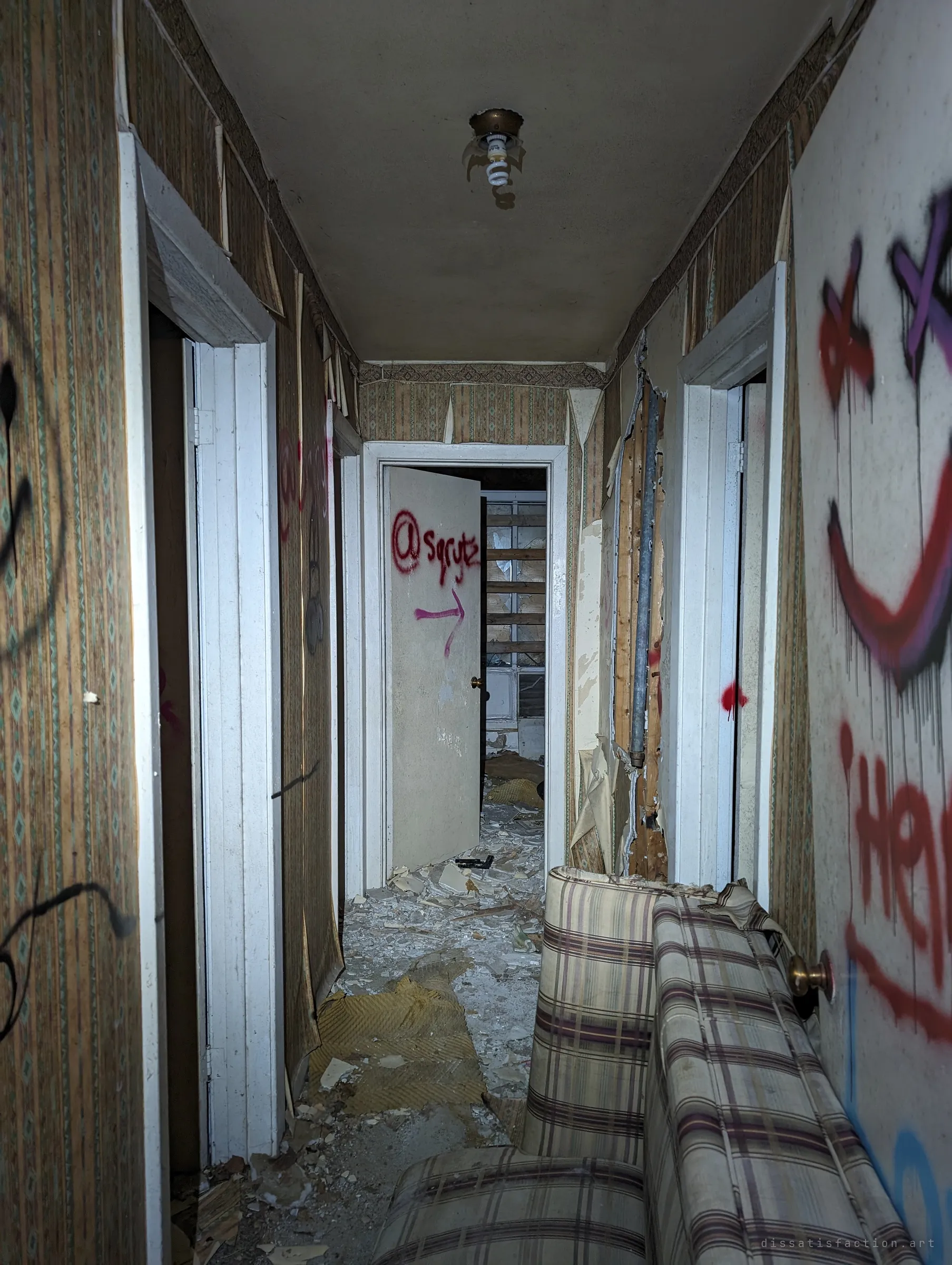 A graffiti-covered corridor littered with plaster and a plaid couch, with doors opening onto dark rooms. At the end an open door is tagged with an arrow pointing onward.