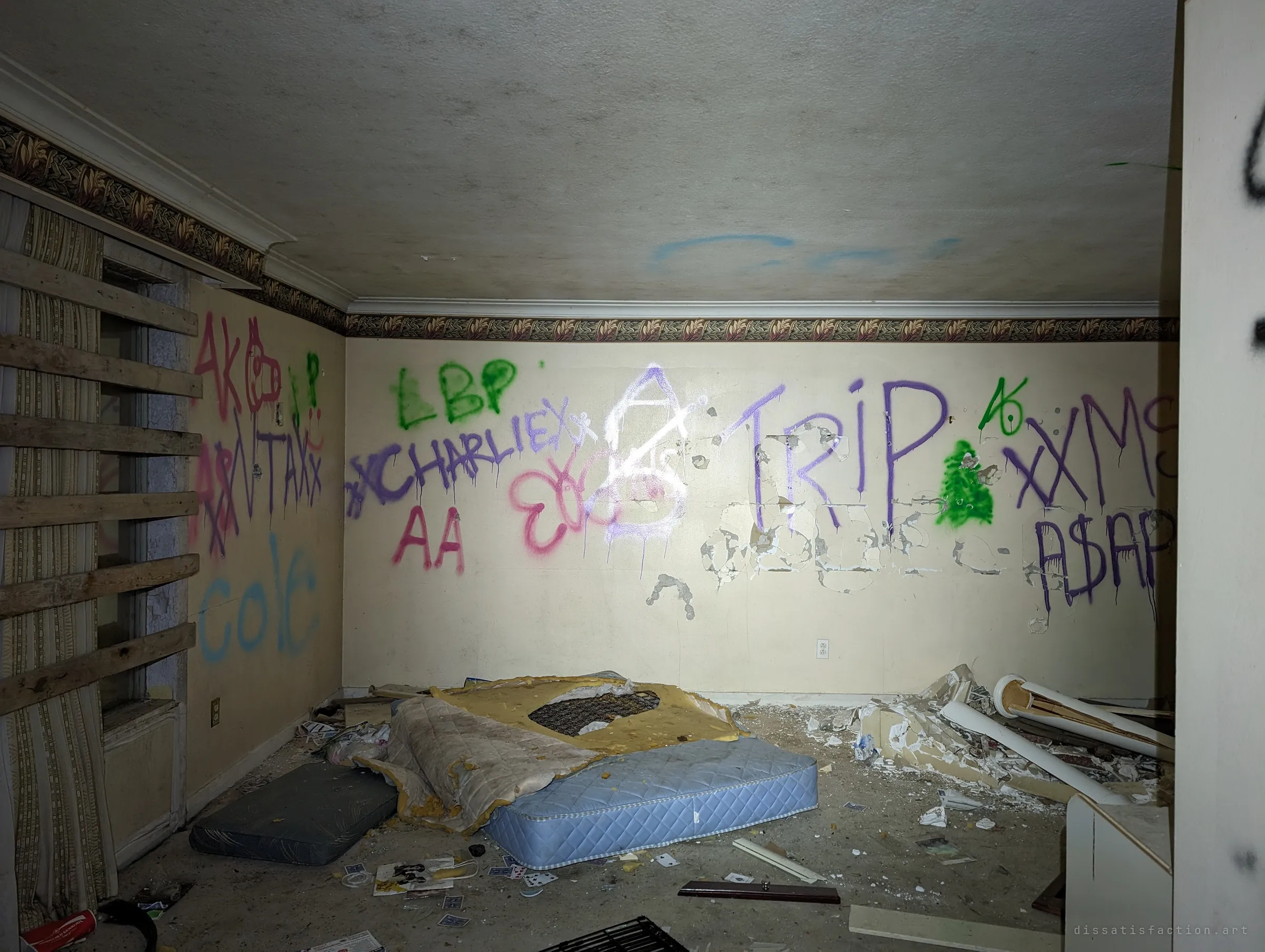 A bedroom with every wall covered in layered tags, bare mattresses and playing cards scattered across the floor. The boarded window at left keeps it dark.