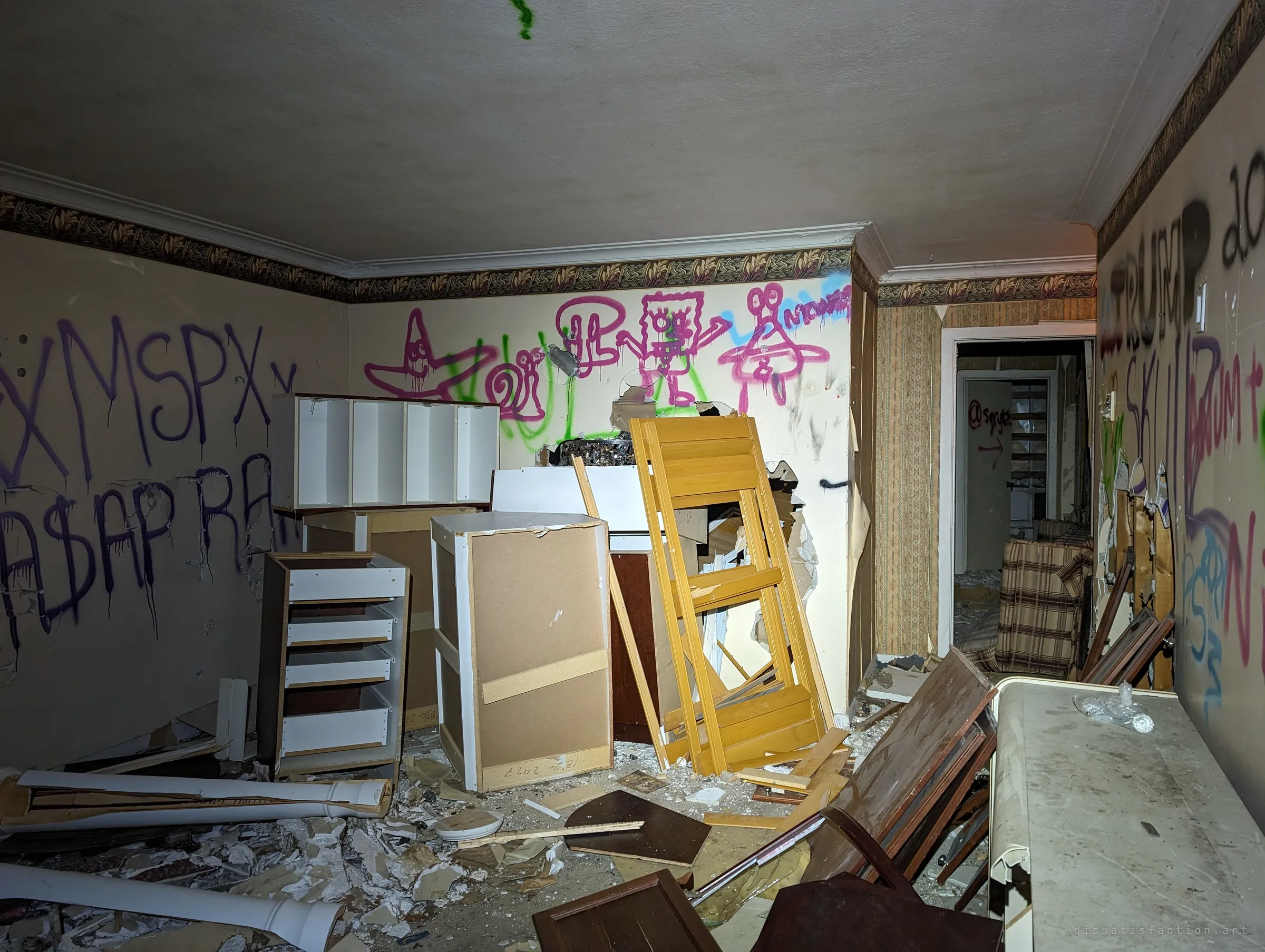 A room crammed with pulled-out kitchen cabinets, broken frames and cartoon graffiti along the top of the wall. Down the hall, the tagged door and plaid couch from the corridor are visible.