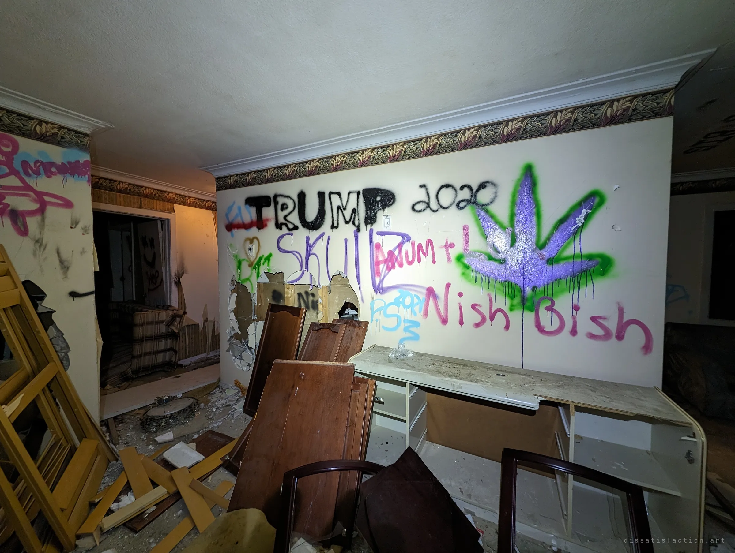 A wide view of a wrecked room where a big painted leaf and a jumble of tags cover the main wall. Cabinet doors and broken furniture are heaped in front of it.