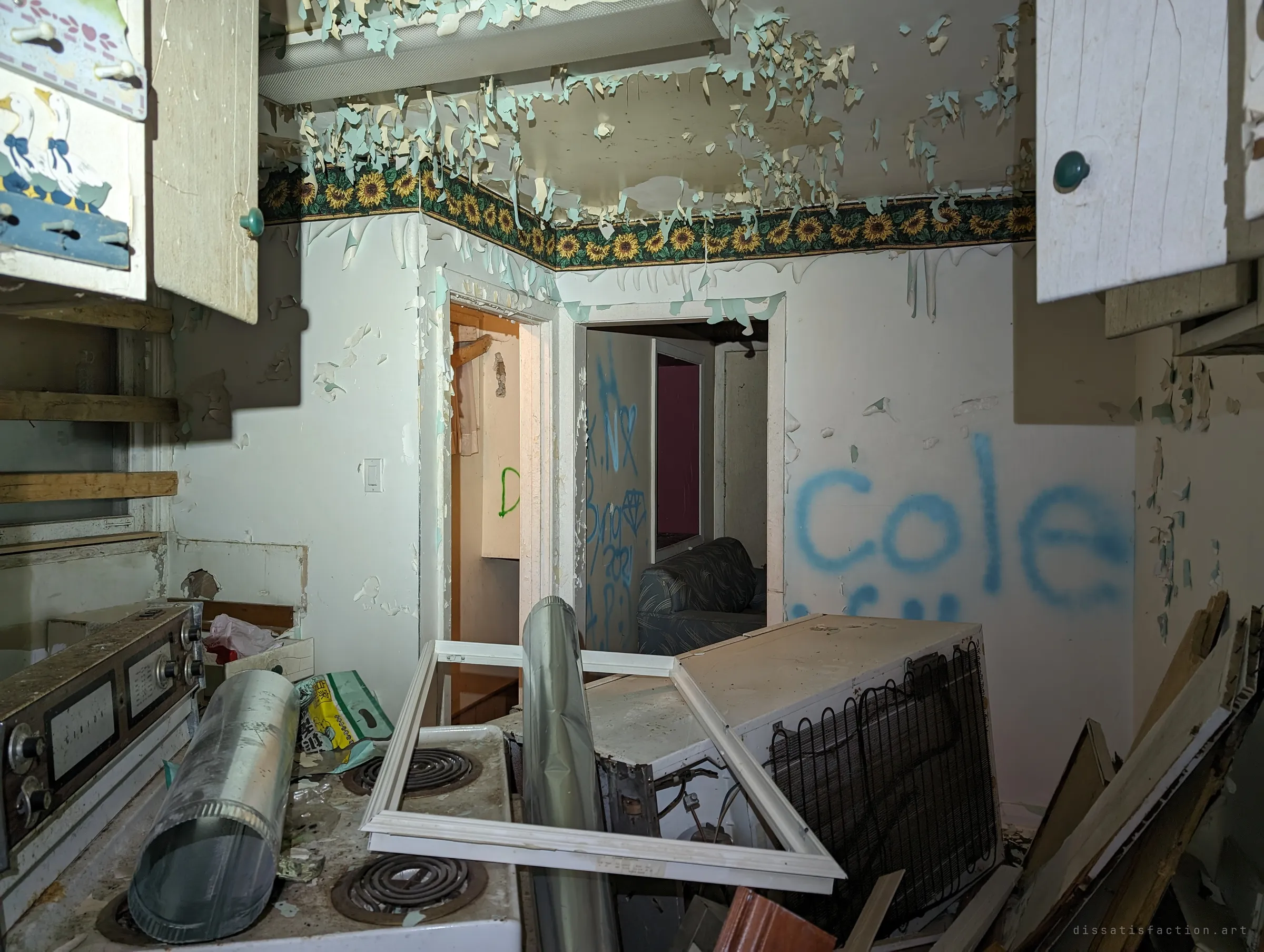 A small kitchen with ceiling paint hanging in curls above a sunflower border, and a stove and fridge buried under ducting and a window frame. Blue tags carry on through the doorways beyond.