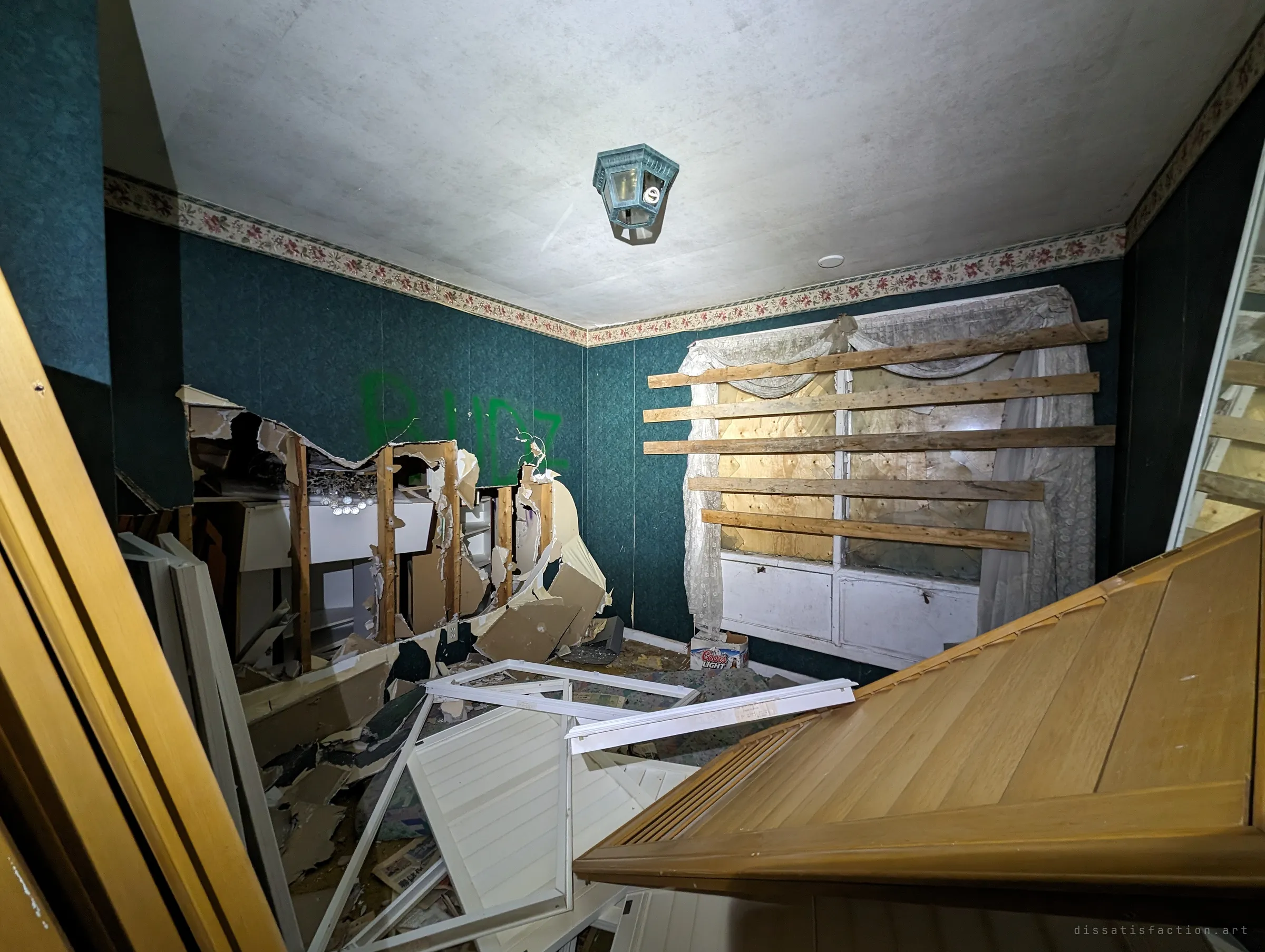 A green-papered room with its wall ripped open to the studs and a boarded window hung with lace curtains. Broken doors and frames lie tipped across the floor.
