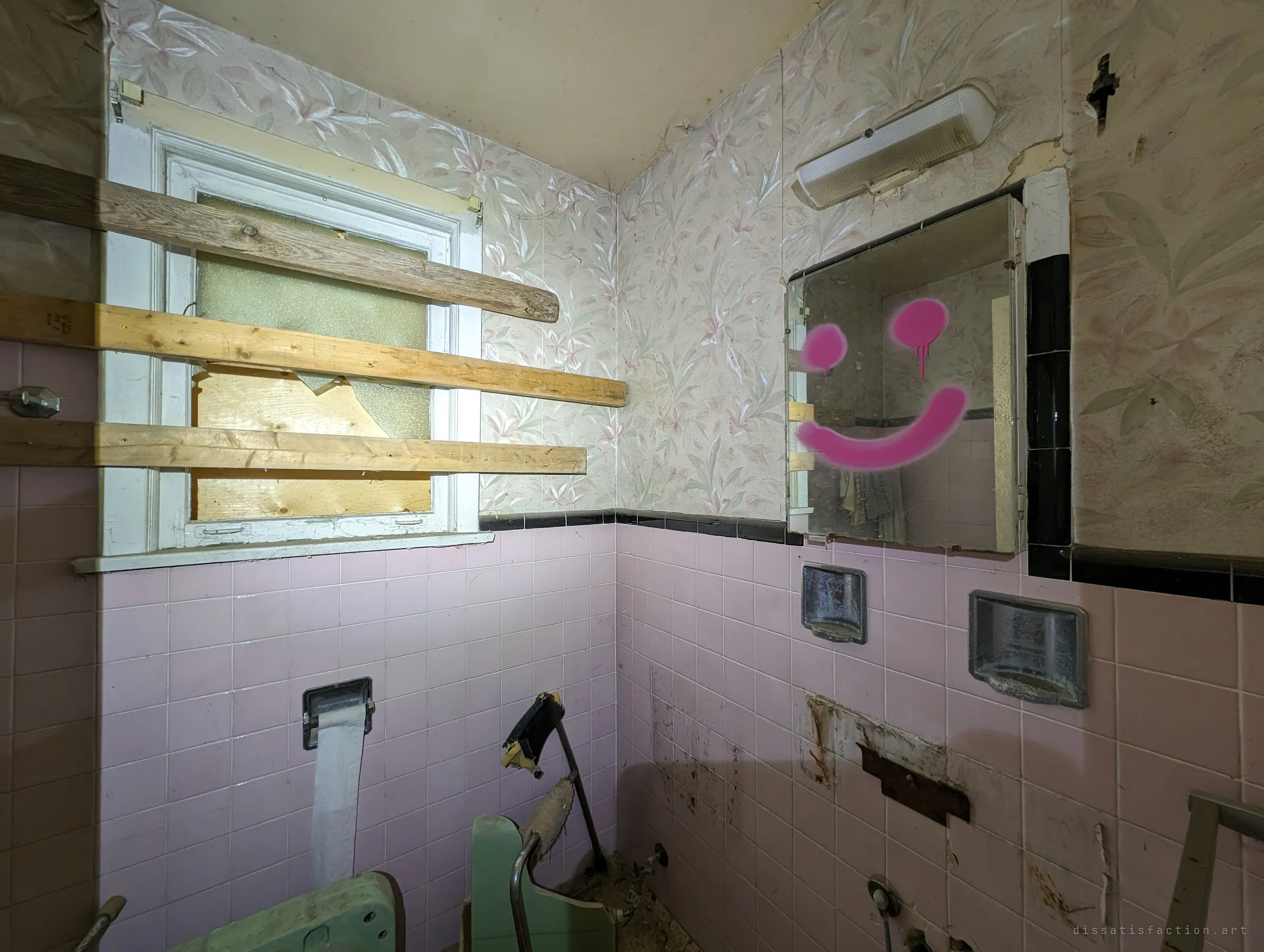 A pink-tiled bathroom with a boarded window and a medicine-cabinet mirror spray-painted with a dripping smiley face. The fixtures are torn out and only a length of toilet paper still hangs.
