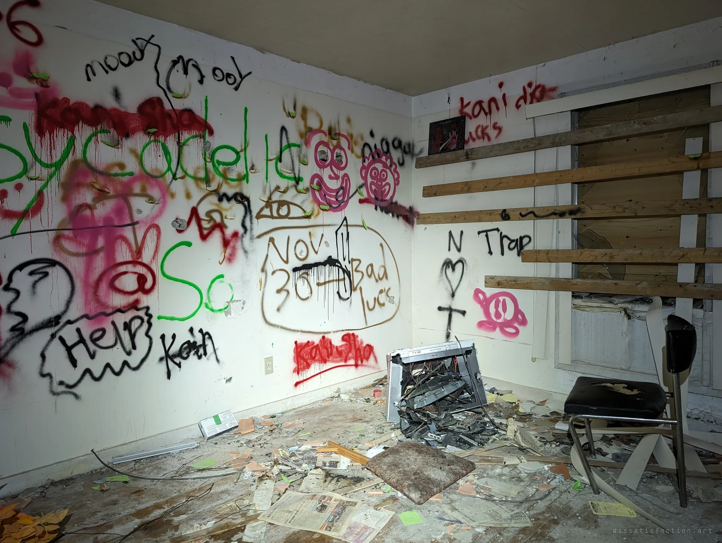 A corner room almost fully covered in scrawled graffiti, faces and messages, with a smashed appliance and a chair among old newspapers. Planks are nailed across the window.