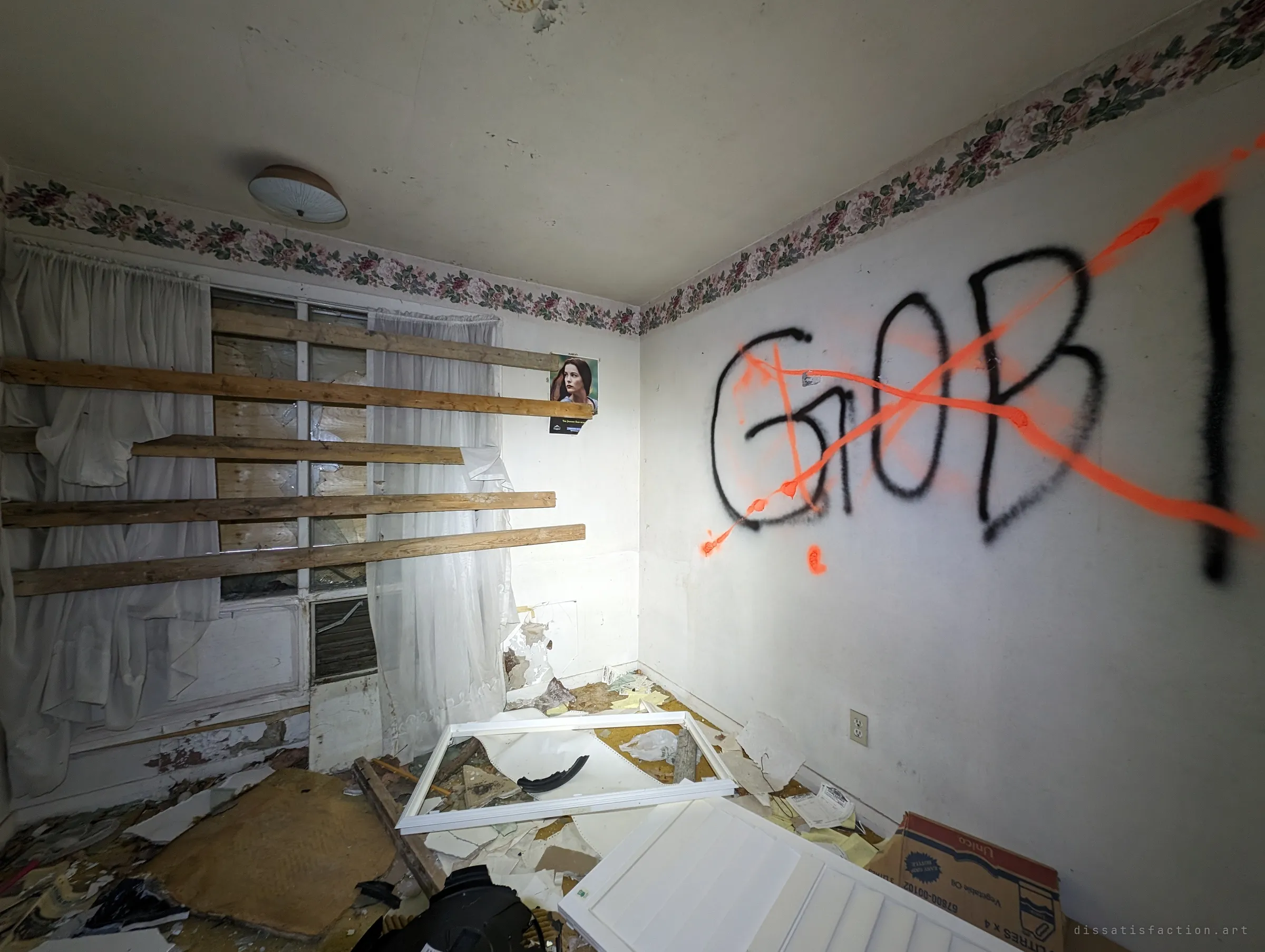 A small bedroom with a large crossed-out tag on the wall, torn curtains and planks over the window. A fallen window frame and shutter lie in the debris on the floor.
