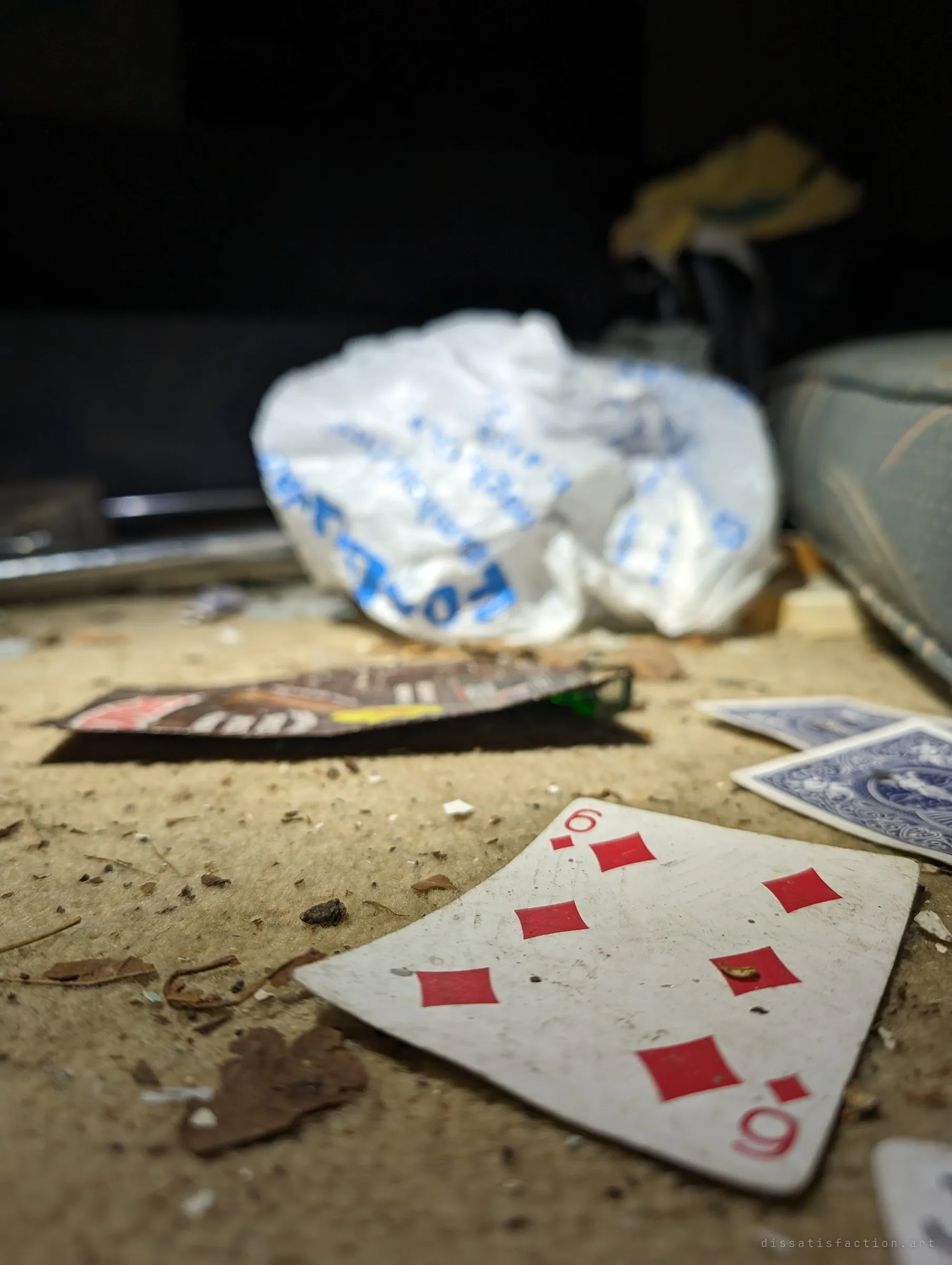 A close, low view of a six of diamonds and other playing cards in the dust, with a crumpled plastic bag blurred behind. It is a quiet trace of whoever passed the time here.