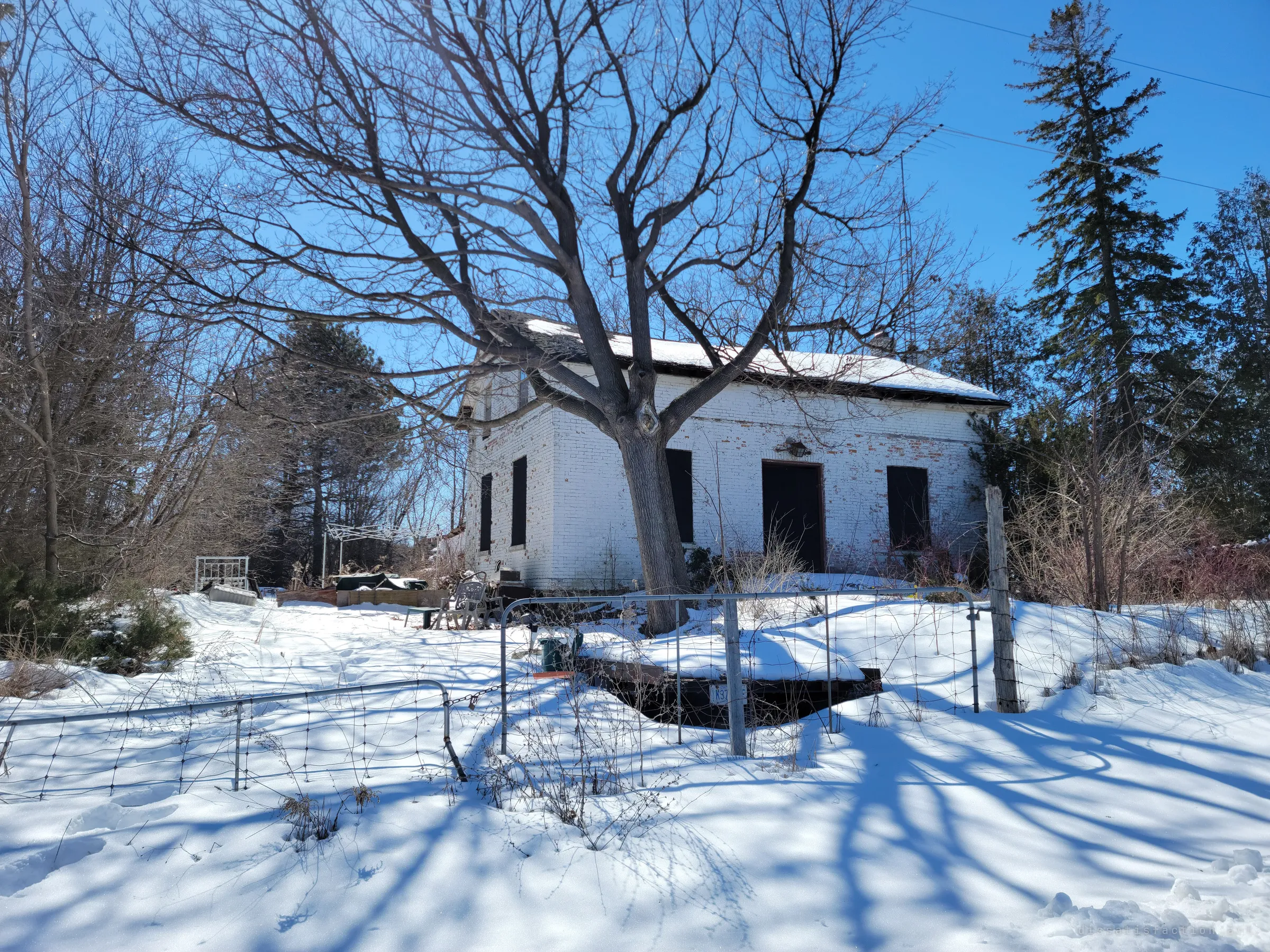 A white-painted brick farmhouse sits behind a bare tree and a sagging wire fence, snow drifted deep across the yard. Boarded windows and a failing roofline set up the decay inside.