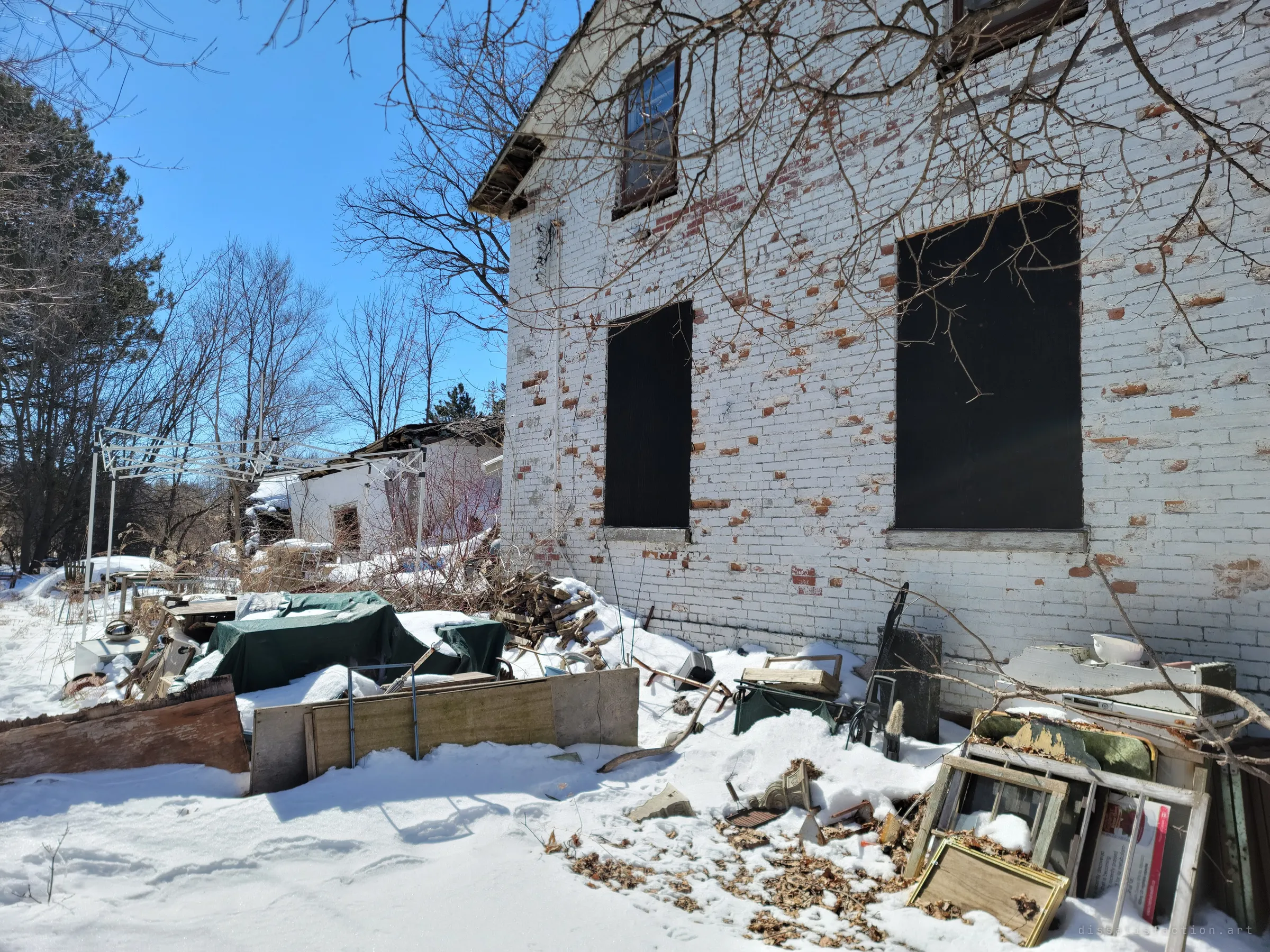 The side wall of the house, its white paint flaking off red brick, with two boarded windows above a yard heaped with tarps, lumber and old window frames. A collapsing outbuilding shows beyond a bent canopy frame.