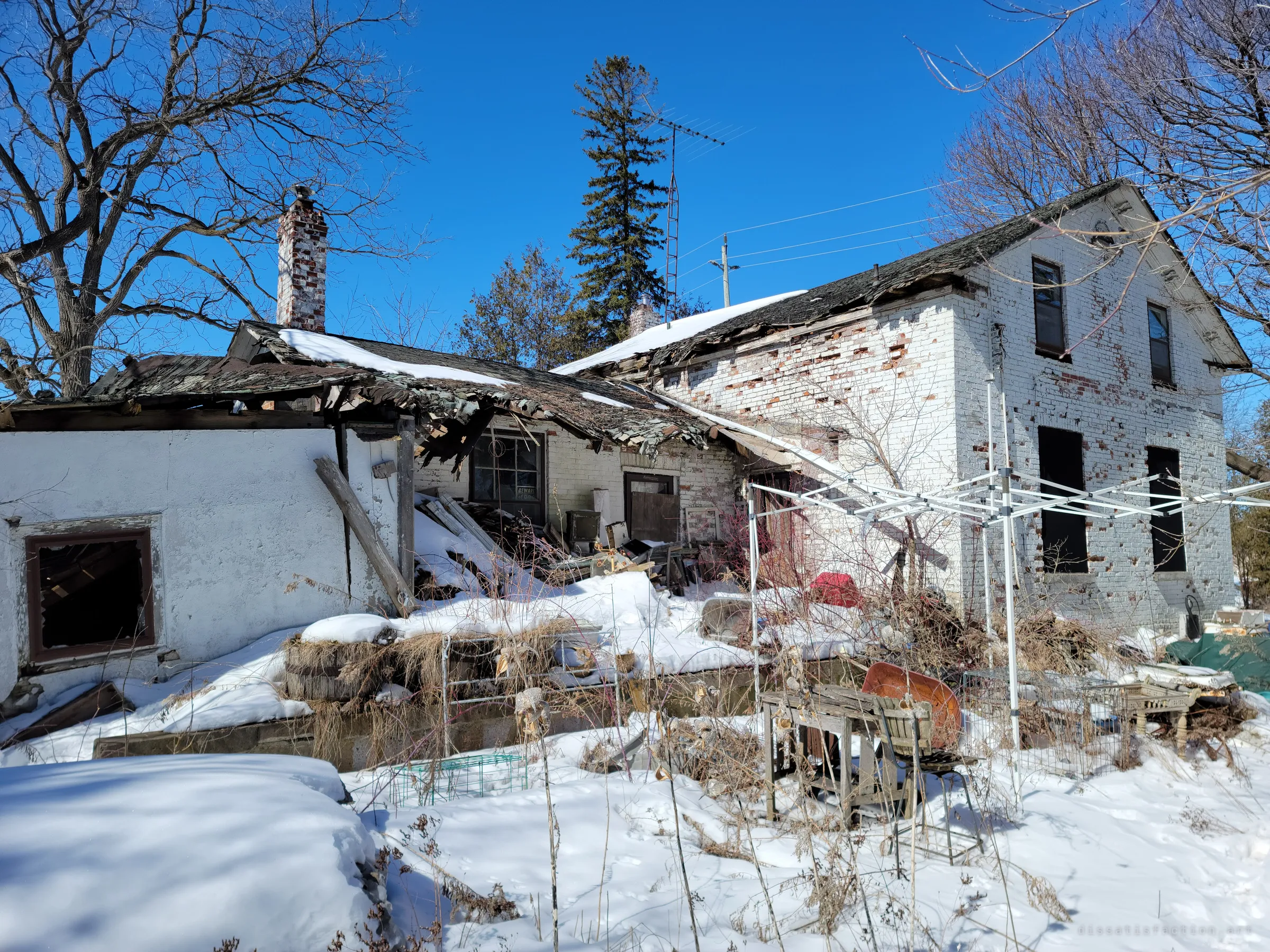 The rear of the house, where a single-storey addition's roof has caved in onto its porch beneath a brick chimney and an old TV antenna. Junk and a rusted wheelbarrow poke out of the snow in front.