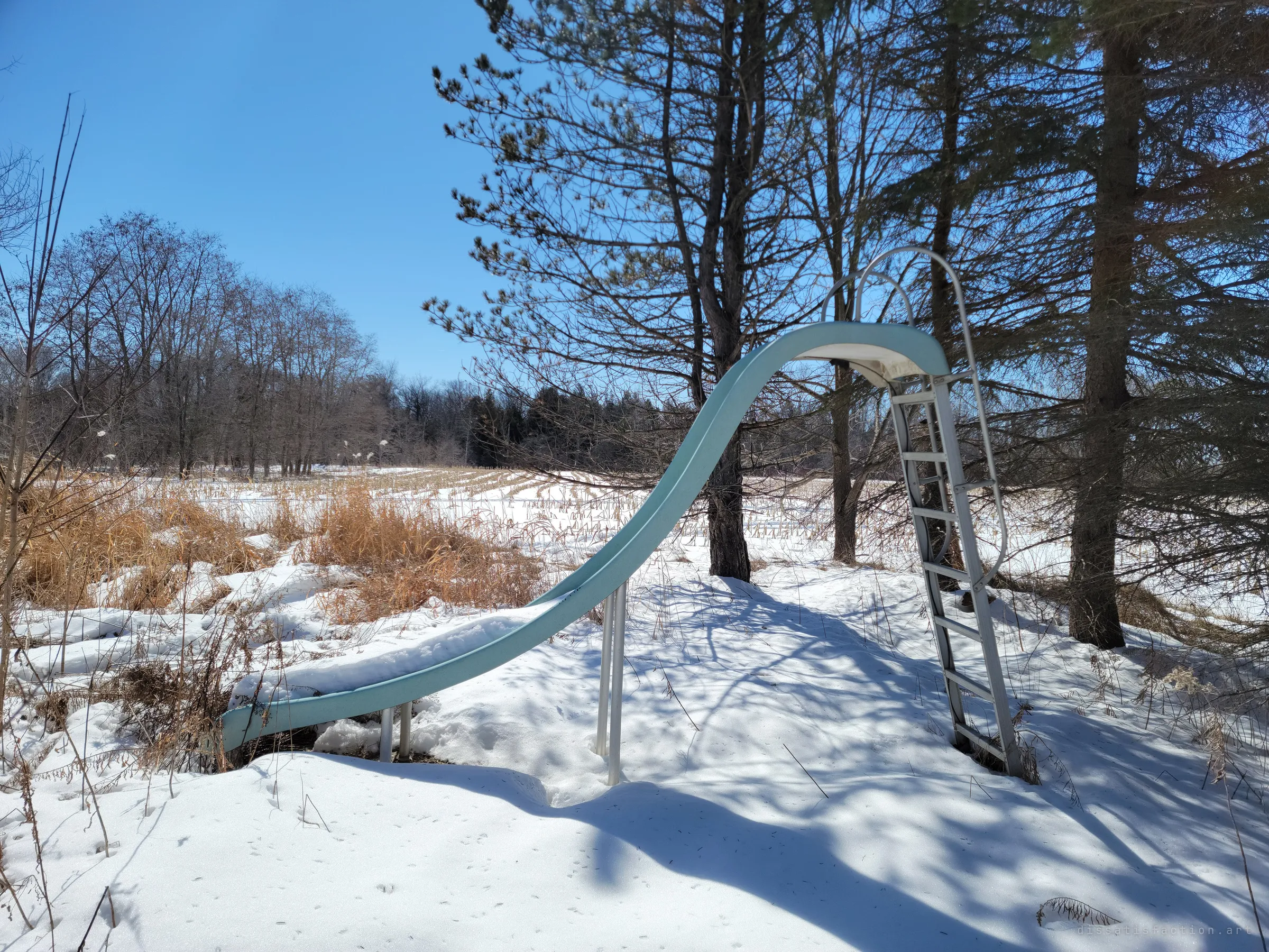 A pale green pool slide stands alone in the snow among pines, with no pool left beneath it. A stubbled field fills the background.