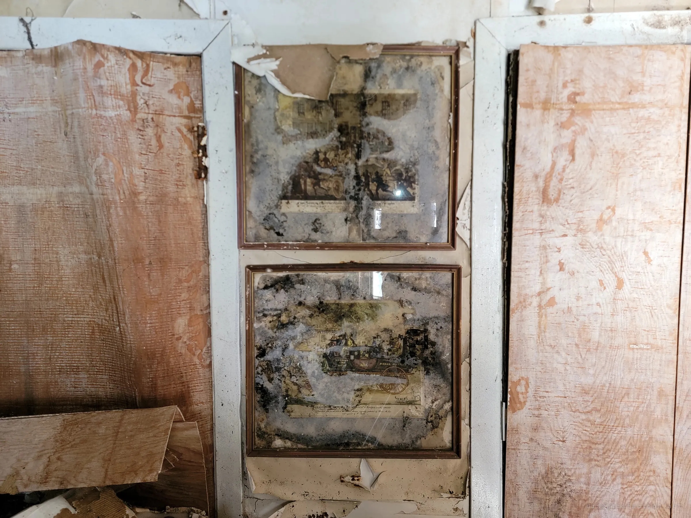 Two framed antique coaching prints hang between panelled doors, their glass blooming with black mould. Damp has eaten through the paper behind them.