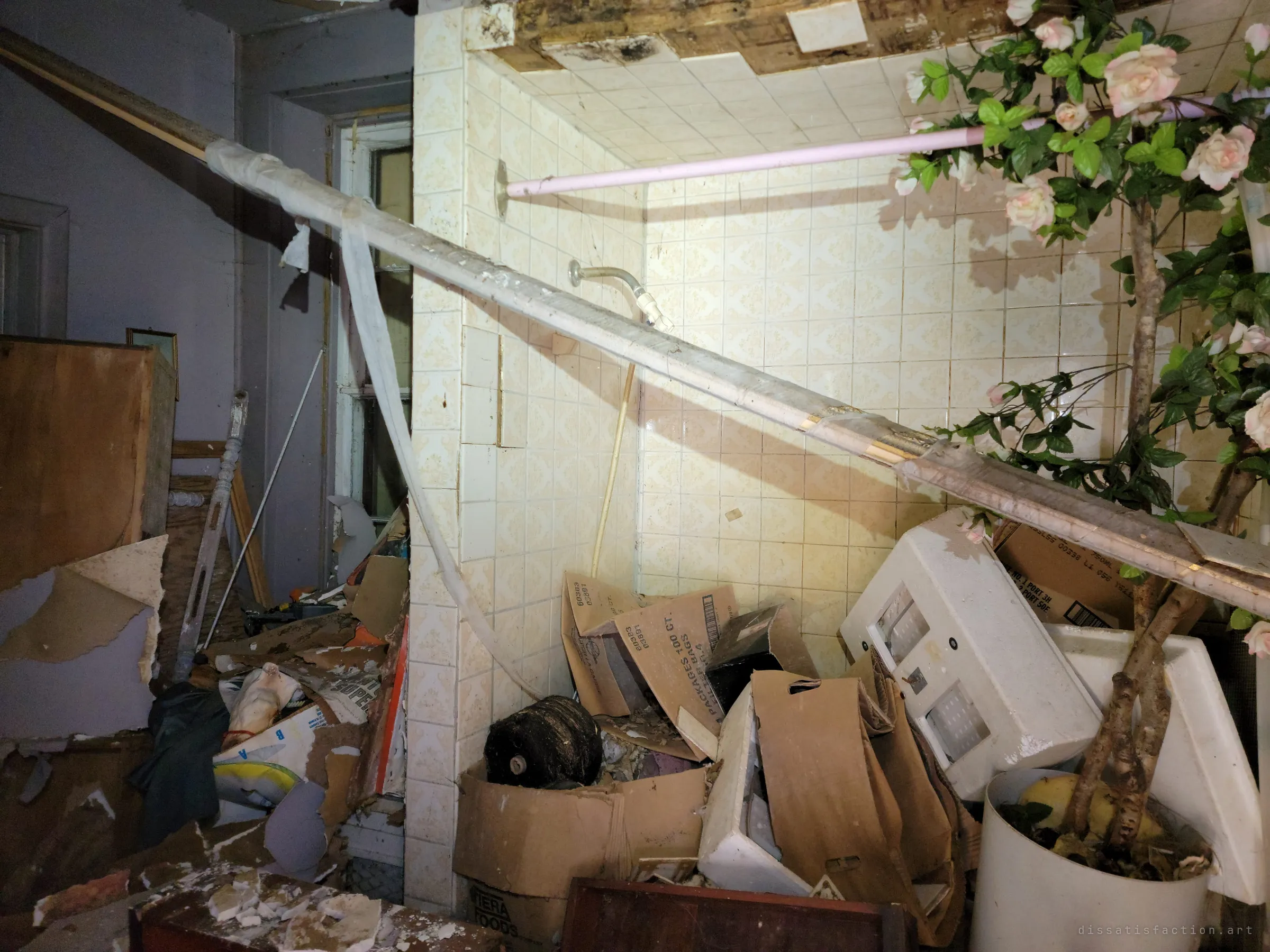 A tiled shower stall packed with cardboard boxes, a toppled cabinet and an artificial rose tree, with a fallen roof beam slanting across it. The pink curtain rod is still in place above the mess.