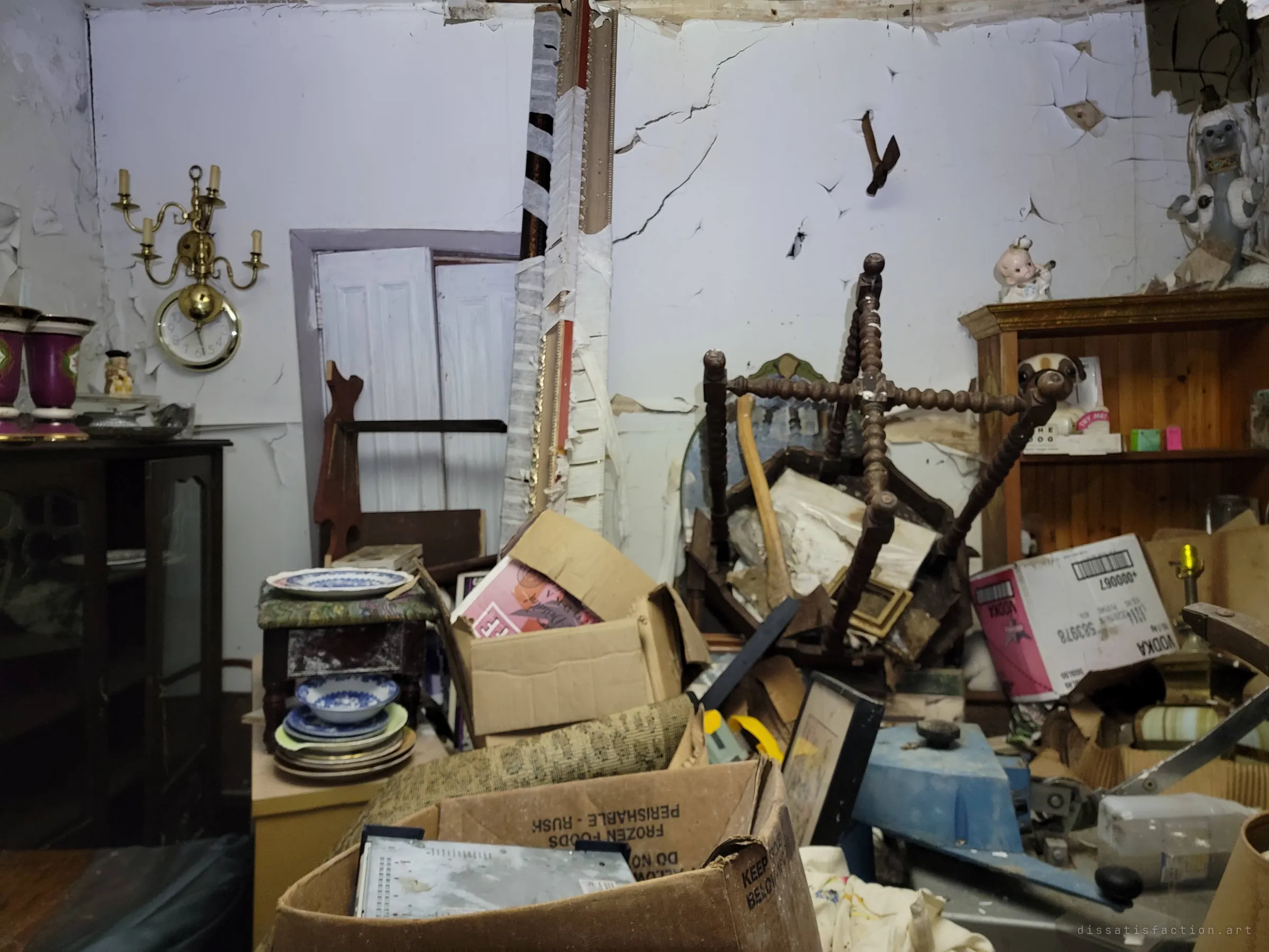A hoarded sitting room piled with boxes, china and an upturned spindle chair beneath a cracked, bulging plaster wall. A brass wall clock and sconce still hang above the clutter, a household frozen mid-collapse.