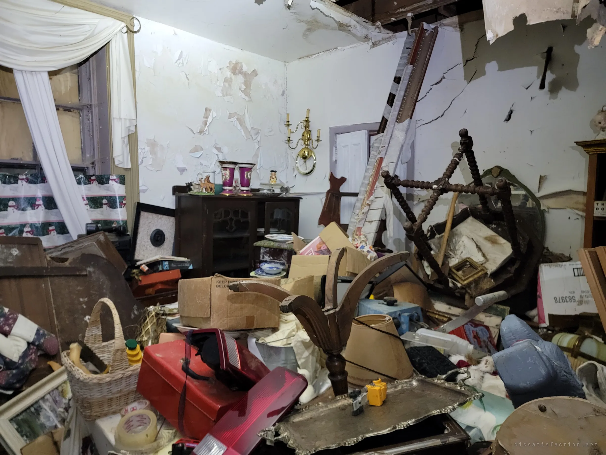 Wider view of the same room: a boarded window draped in a sheer curtain, snowman gift wrap, and furniture buried under boxes and a toppled chair. Peeling paint and a sagging ceiling show how far the house has rotted.