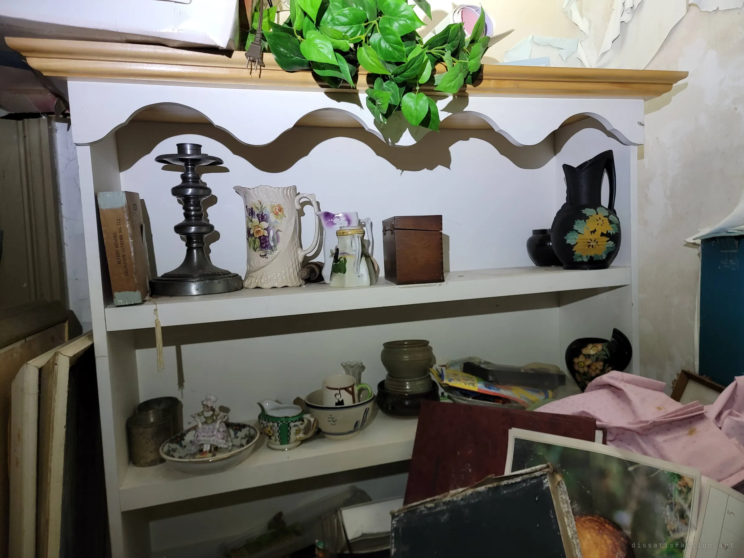 A white hutch still neatly set with painted pitchers, a pewter candlestick and small pottery, under a trailing fake plant. An oddly tidy corner inside the decay.