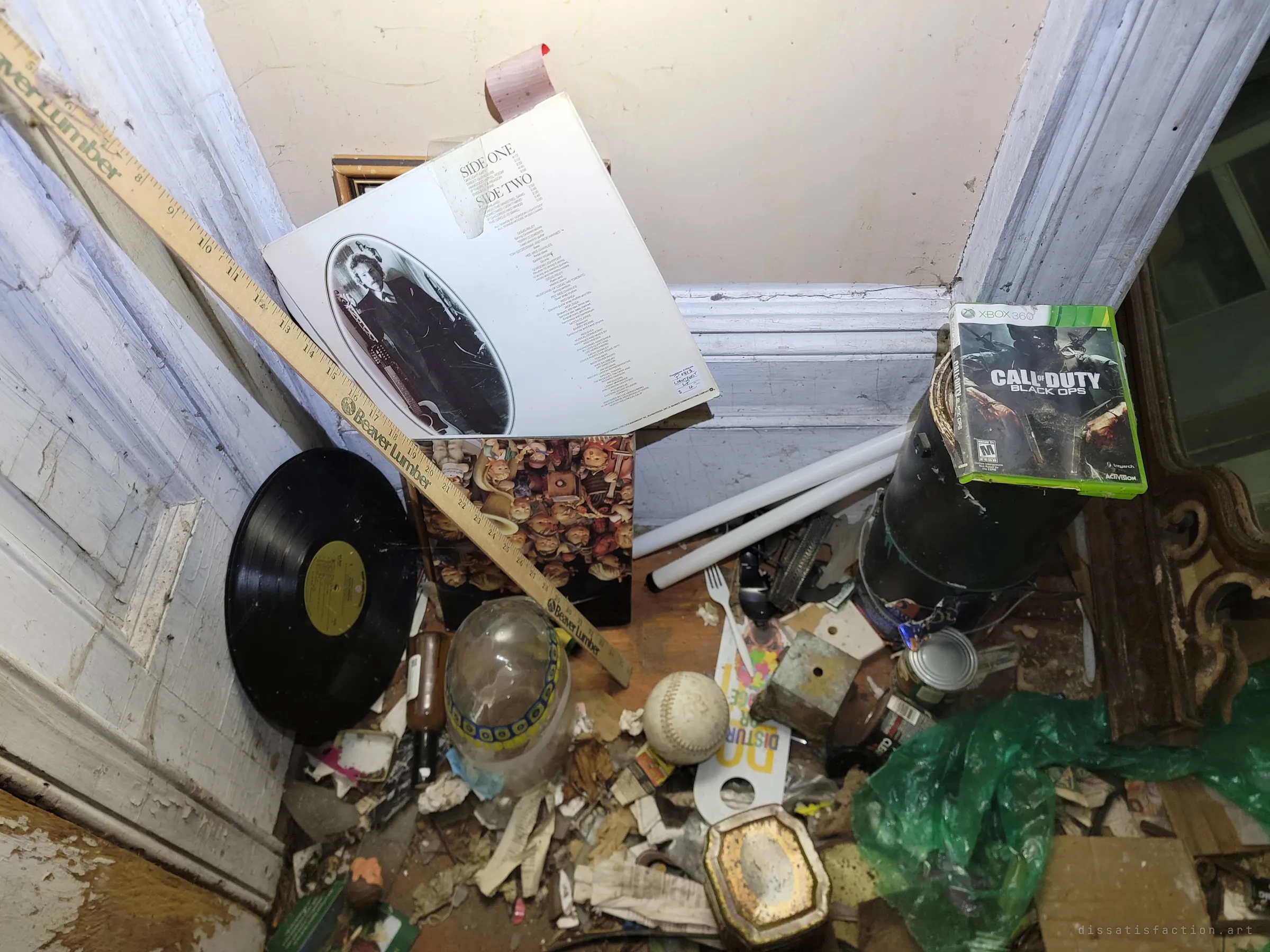 Looking down into a cluttered corner: an old vinyl record and its sleeve, a yardstick, a softball and a video game case among paper scraps and debris. Decades of belongings left in one heap.
