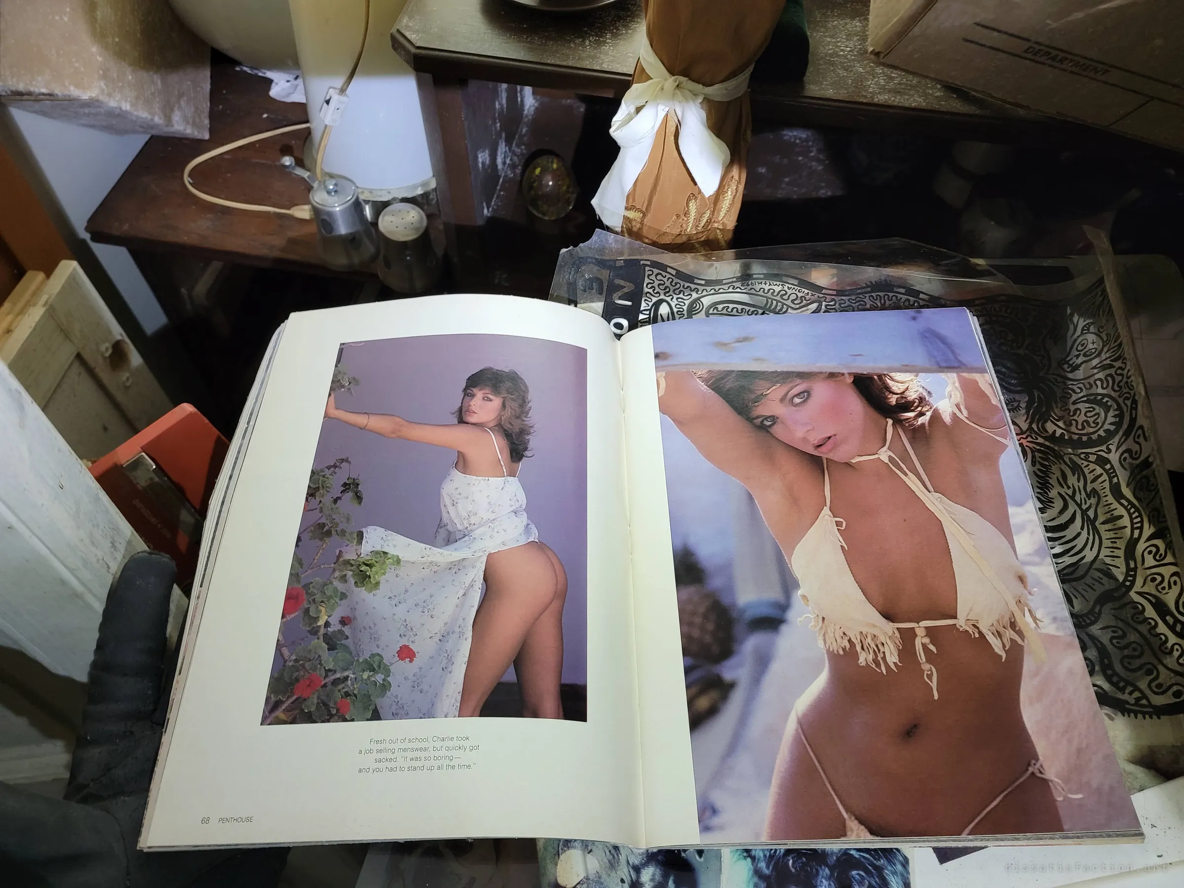 A gloved hand holds open a vintage men's magazine on a cluttered side table beside a lamp and salt shakers. A stray personal relic from the house's past. Note: the spread shows partial nudity.