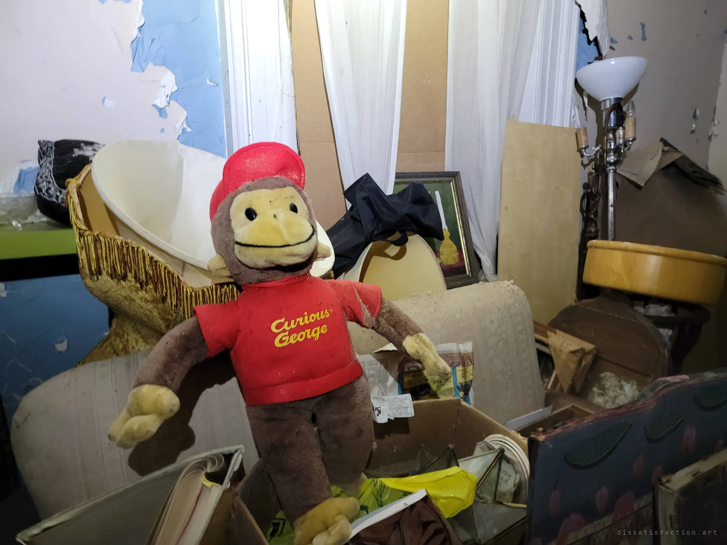 A dusty plush monkey in a red shirt and cap sits propped on a box of lampshades and old newspapers. A child's toy left behind in a room of peeling blue paint.