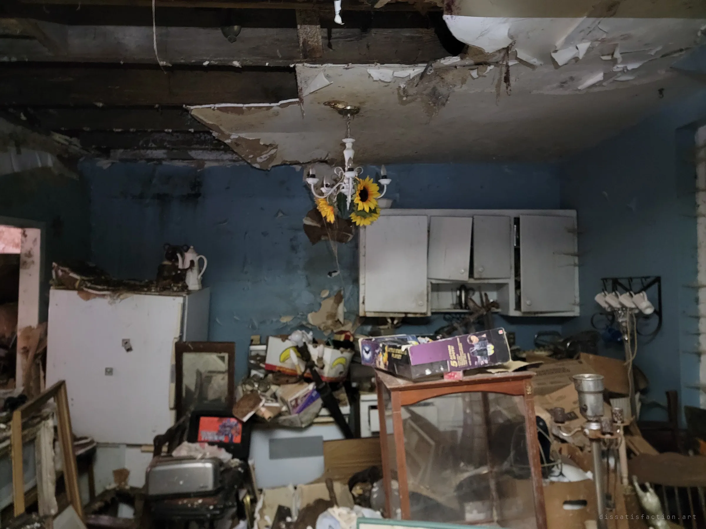 A blue kitchen under a ceiling that has fallen open to the joists, a sunflower-wrapped chandelier hanging over piled furniture and boxes. Water damage has eaten through the plaster above the cabinets.