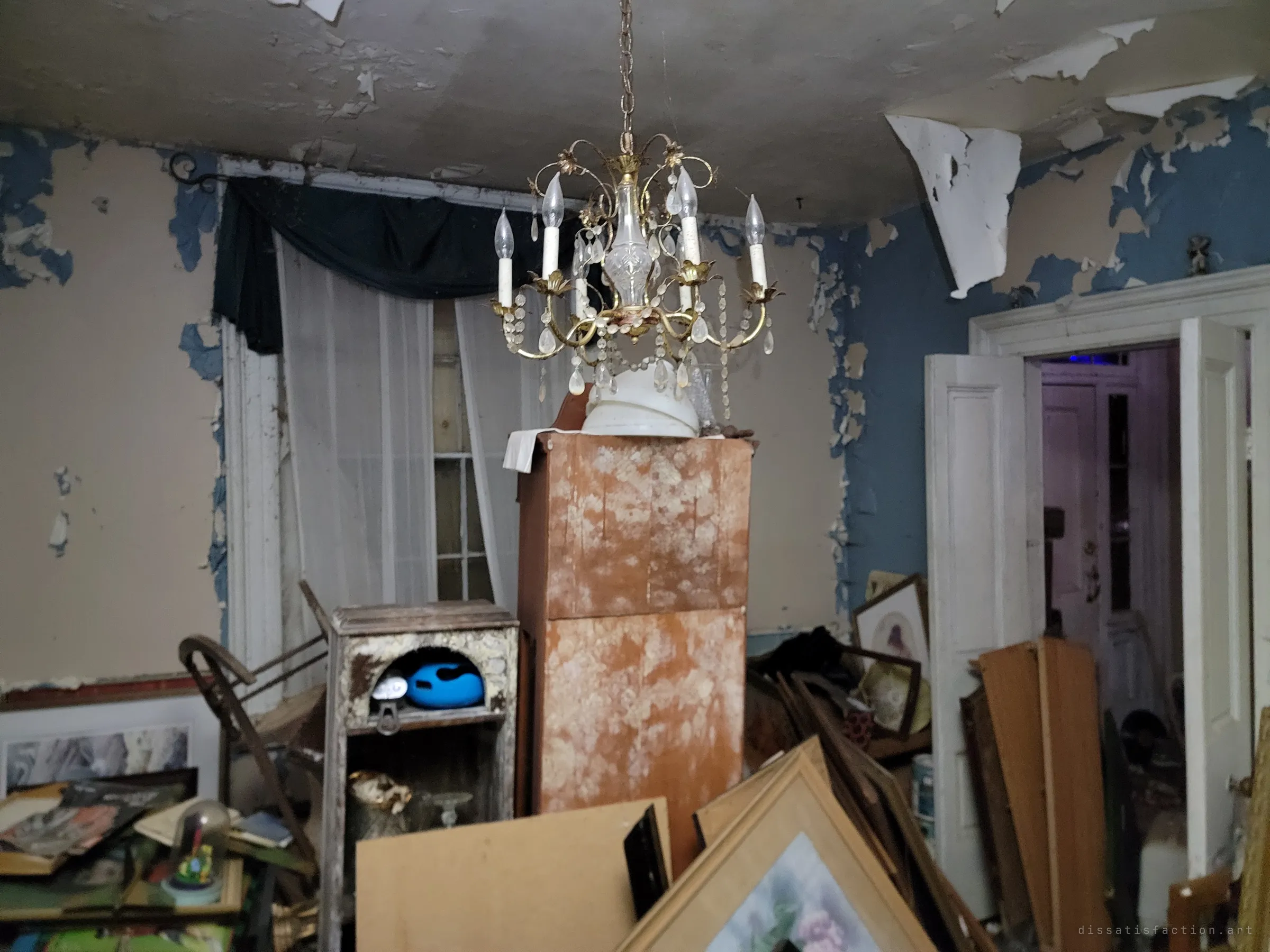 A crystal chandelier still hangs in a front room of peeling blue and beige paint, above stacked frames and a mould-spotted cabinet. Sheer curtains cover the window, and a doorway leads deeper into the house.