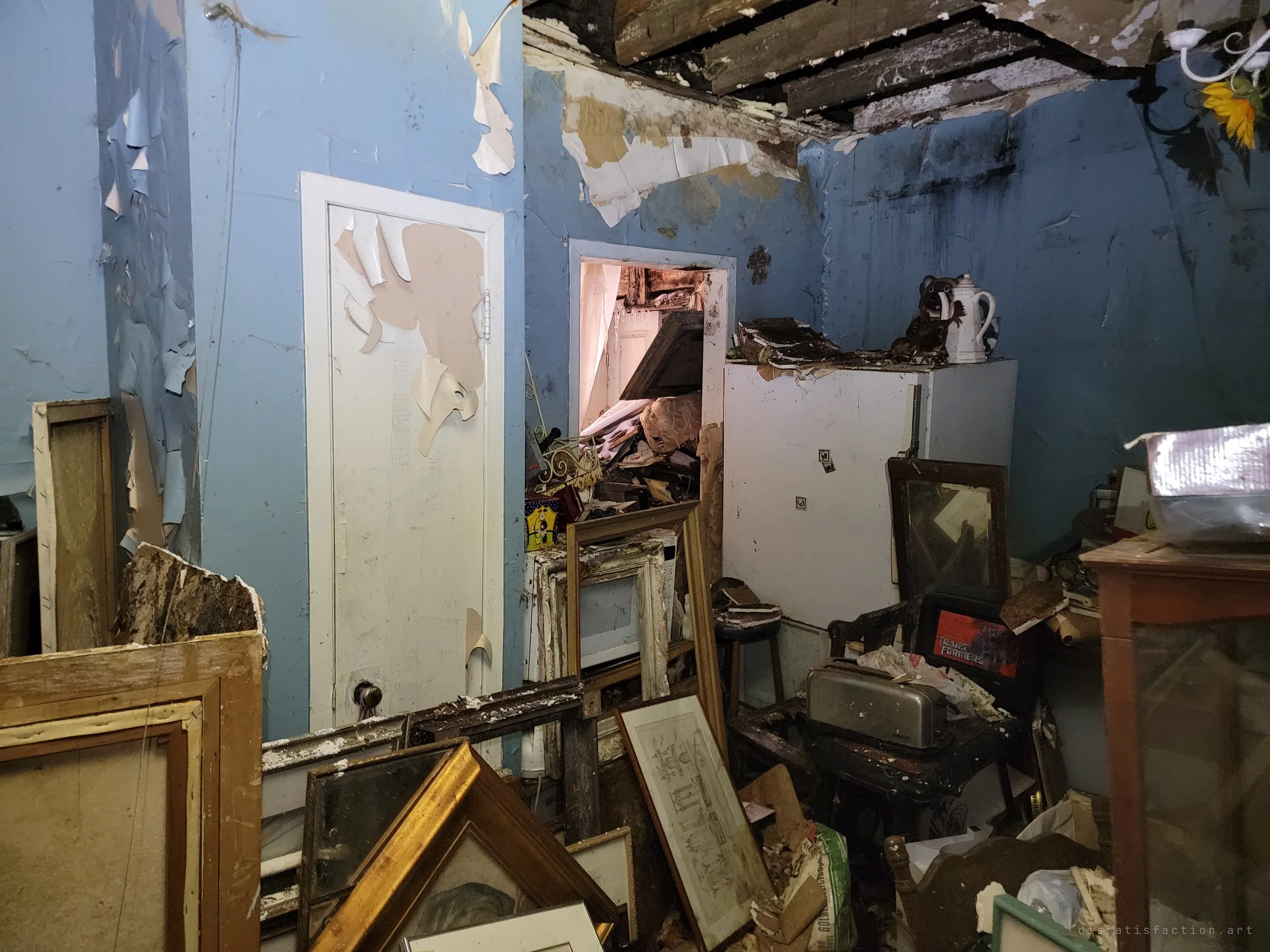 The other side of the blue kitchen: a closed door, a fridge topped with a teapot, and a doorway packed with collapsed debris. Picture frames and a toaster crowd the floor under the broken ceiling.