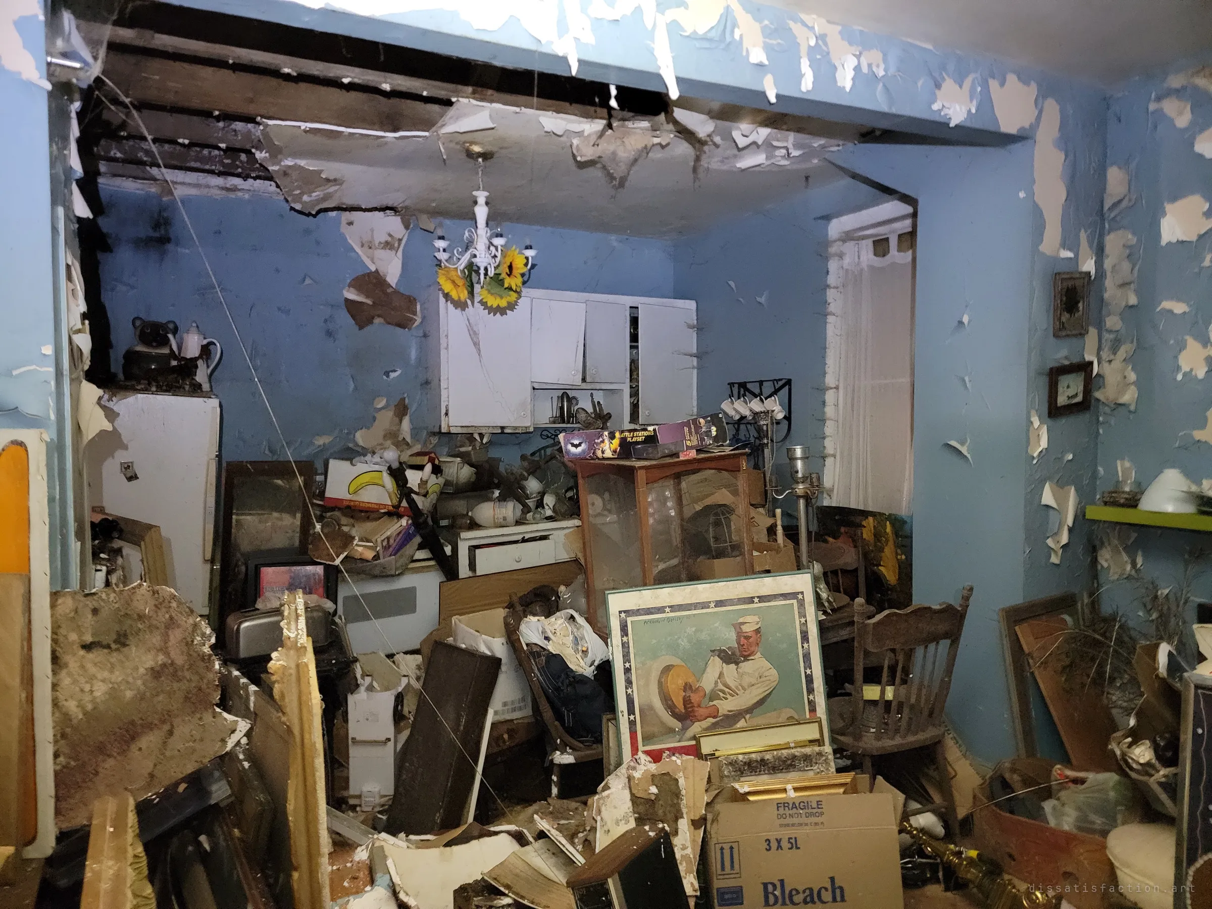 A sky-blue kitchen buried in hoarded belongings under a half-collapsed ceiling, a white chandelier with plastic sunflowers still hanging over the wreckage. The intact decor against exposed joists shows how suddenly the home was left to rot.