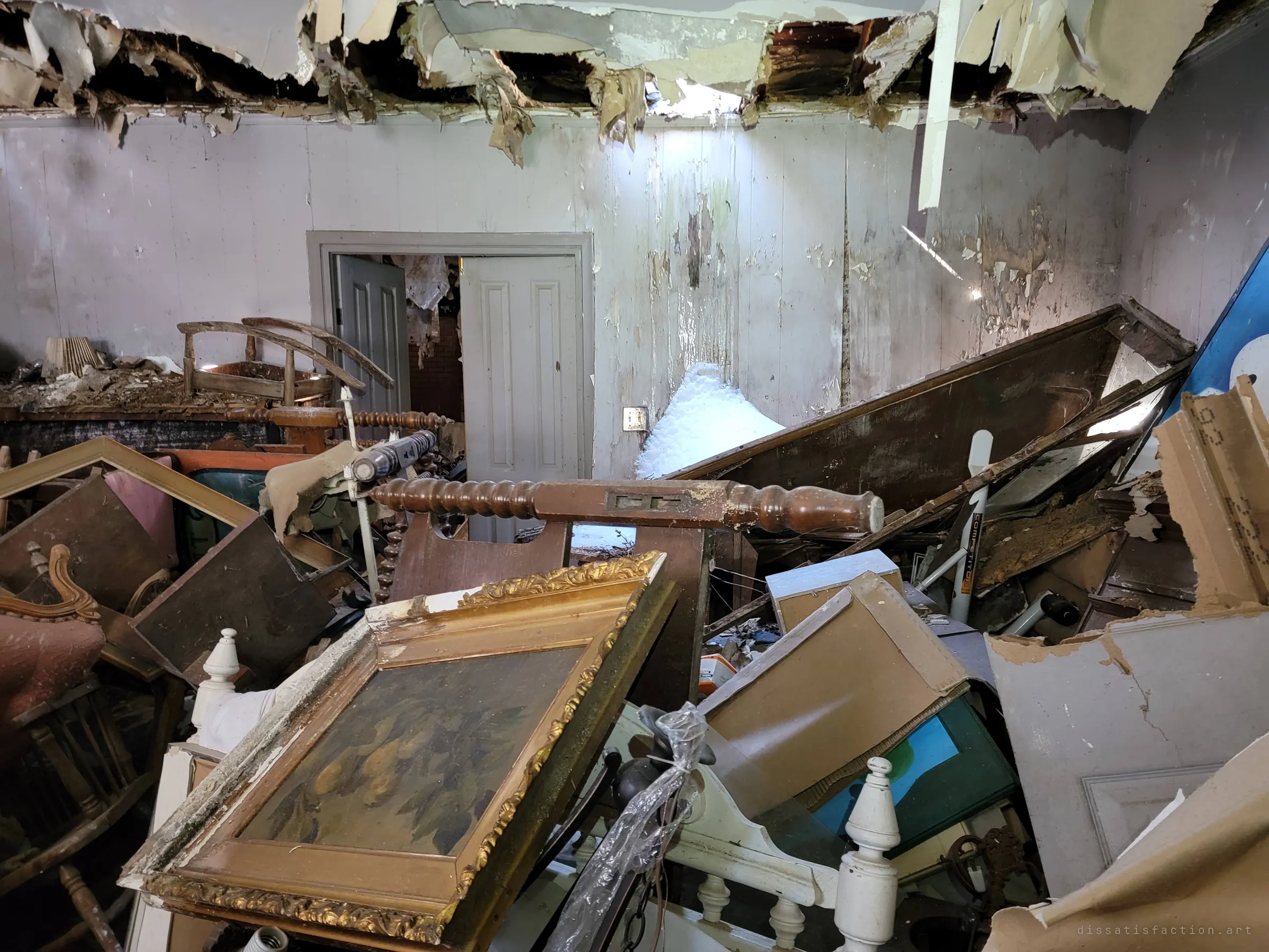 A room crushed by fallen ceiling boards, with spindle bed rails, gilt picture frames and furniture heaped under a torn-open roof letting in daylight and snow. It shows the structure actively giving way over the owner's possessions.