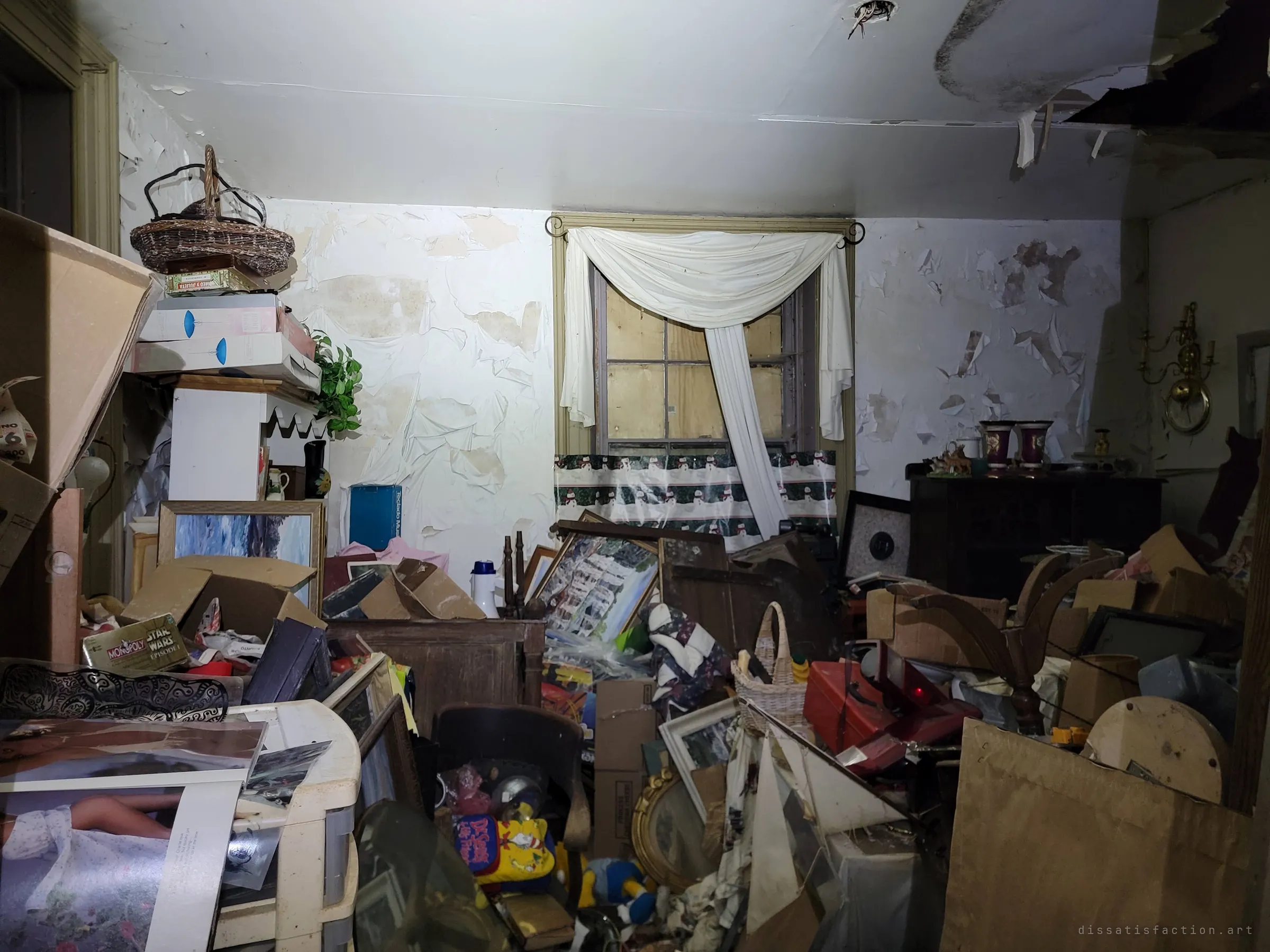 A dim room piled to chest height with boxes, board games, framed prints and toys beneath a boarded-up window draped with a white curtain. The layers of clutter hint at decades of accumulated life.