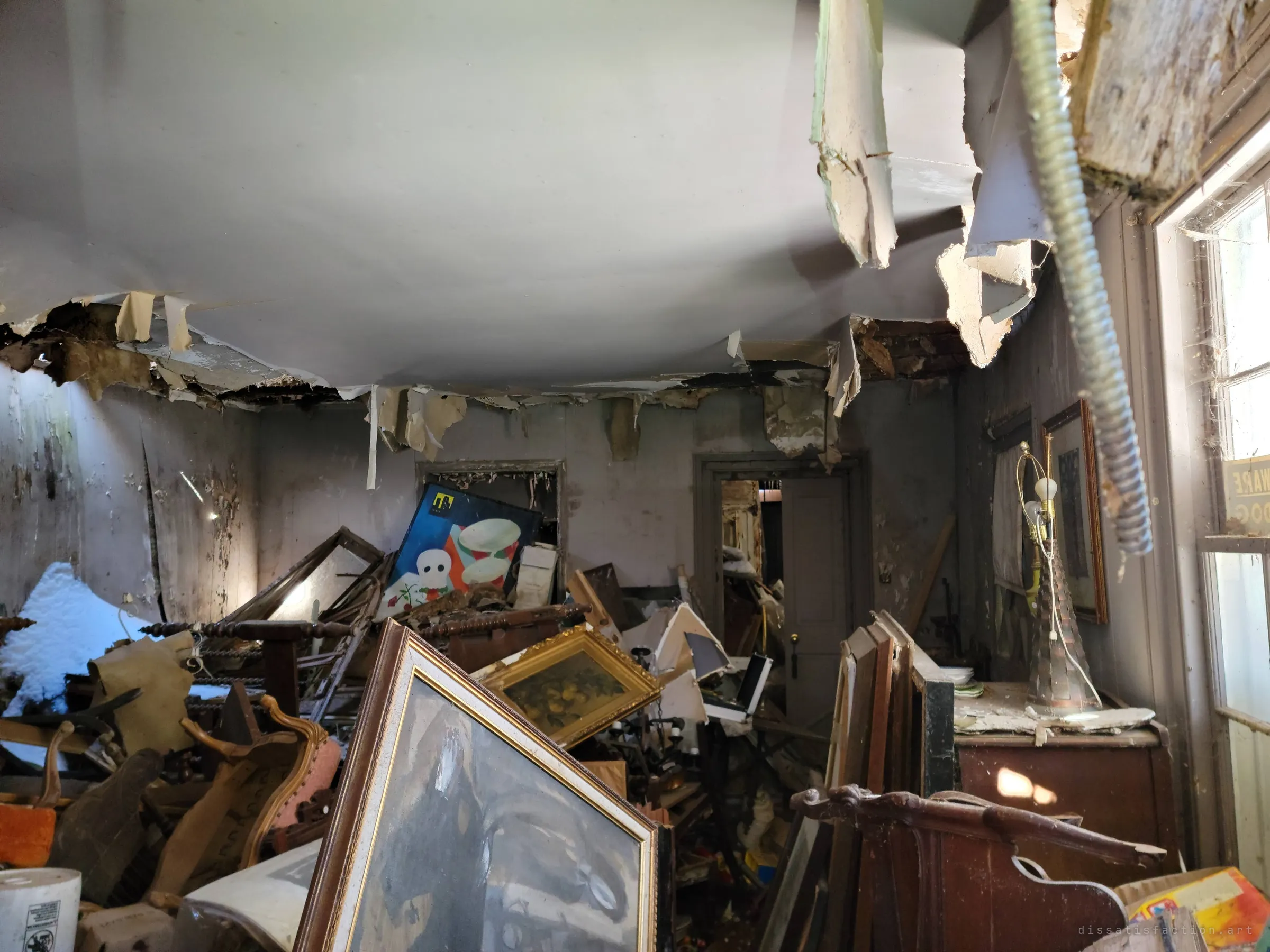 Sagging ceiling panels and dangling conduit hang over a room jammed with frames, chairs and artwork, sunlight slanting in from a side window. The drooping ceiling looks ready to come down on everything below.