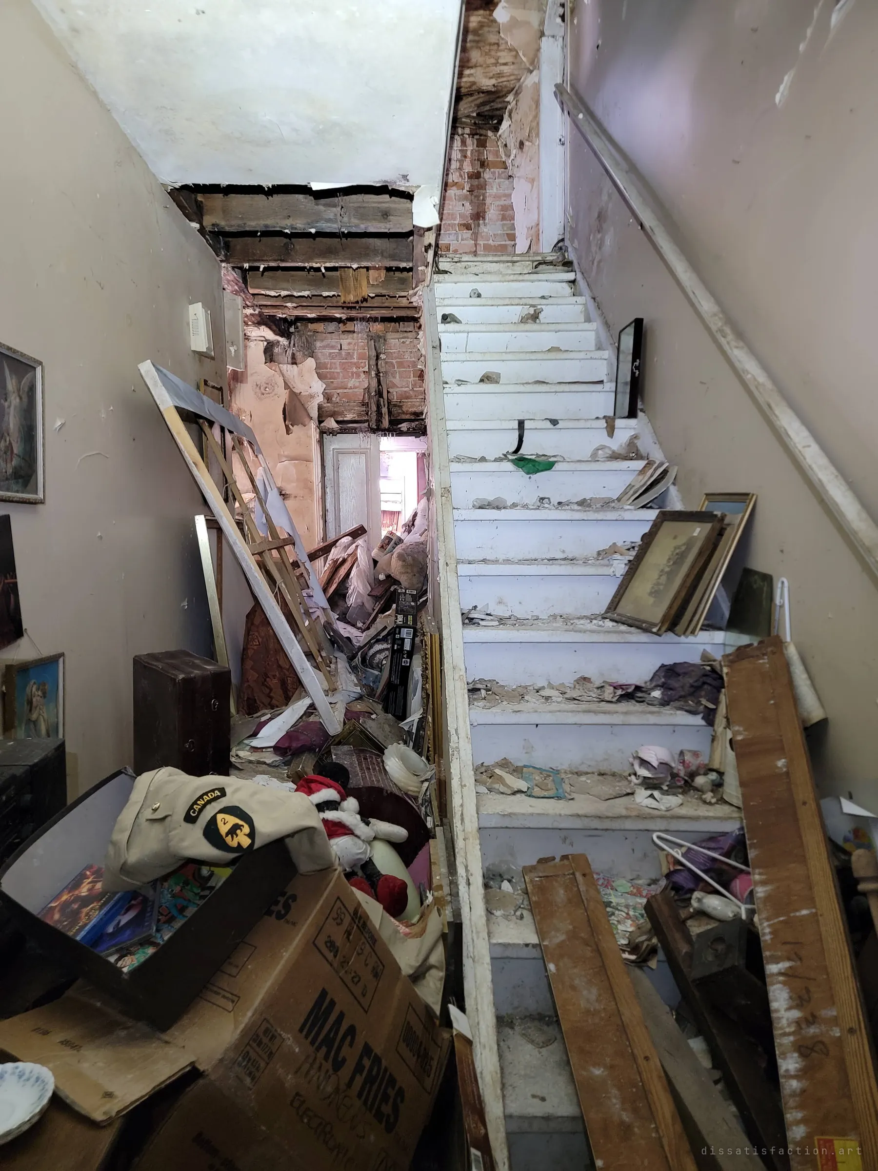 A narrow staircase strewn with plaster and fallen frames rises past a hallway opened up to bare brick and joists, with an old army cap resting on a cardboard box at the bottom. The stair is the route to the upper floor that is coming apart.