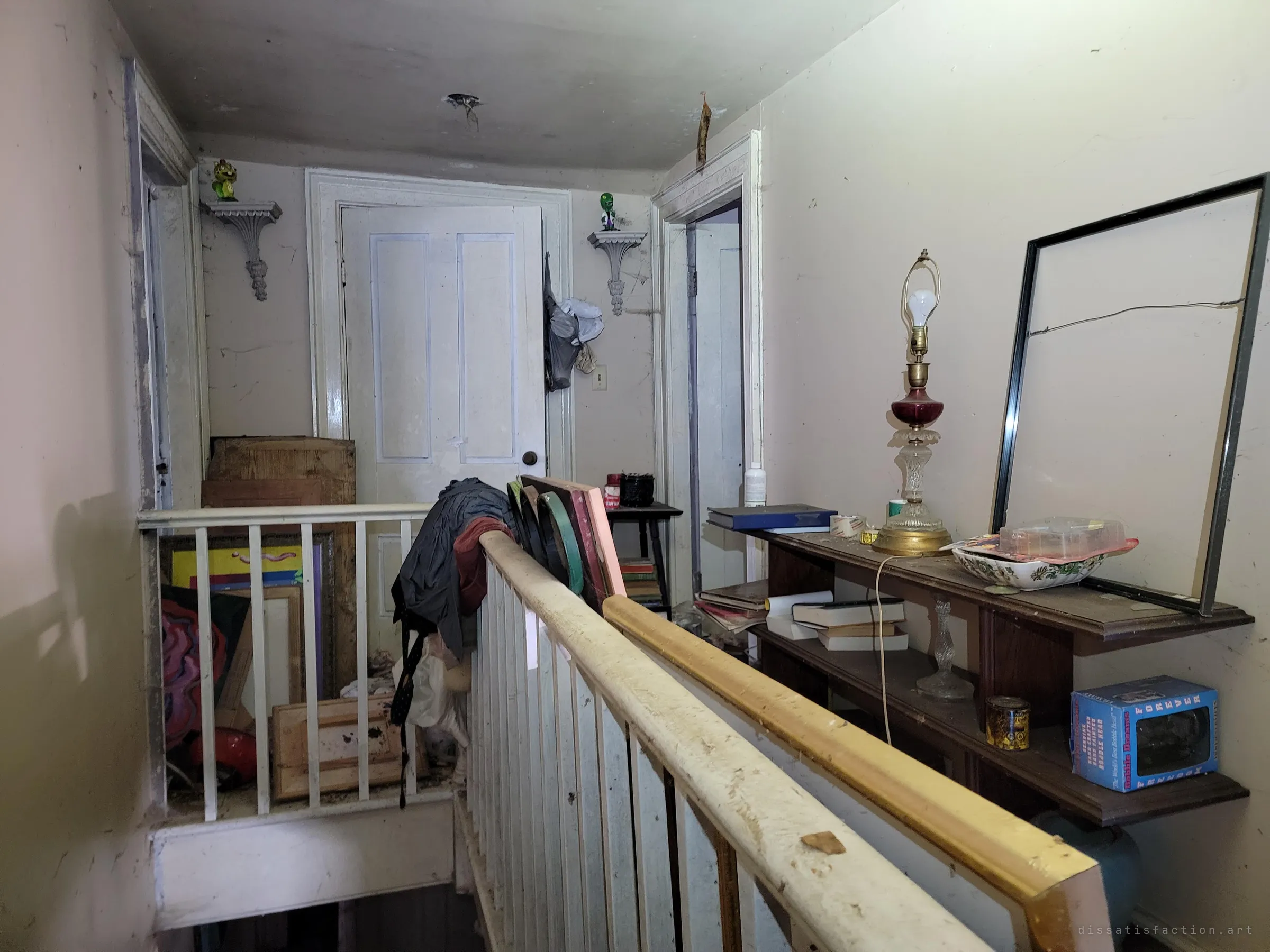 An upstairs landing with a dusty banister, panelled doors and a shelf holding a ruby glass lamp and old books, oddly tidy compared to the floors below. The railing is the only way around to these rooms.