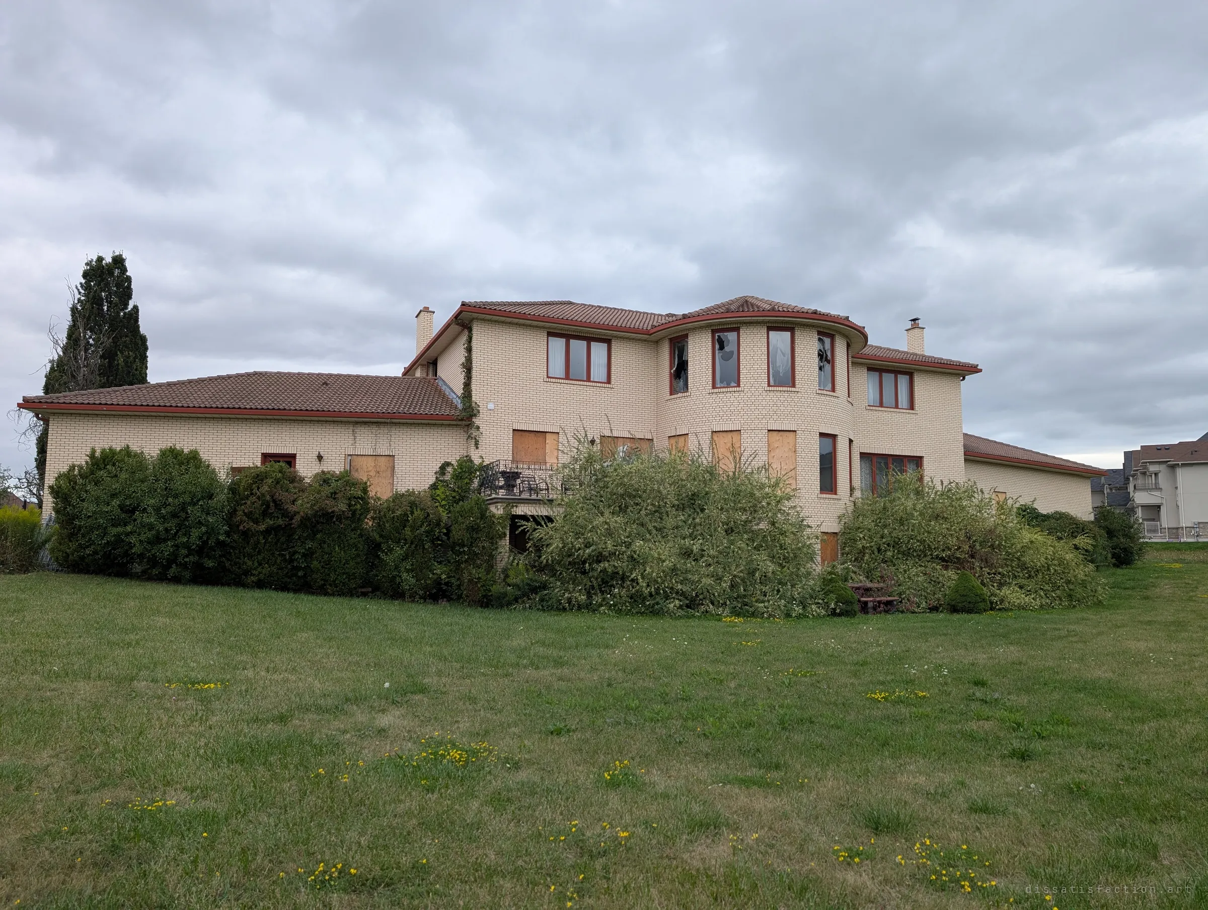 The house seen from an angle across the unmown lawn, its turret windows smashed and the ground floor boarded behind wild bushes. It sits right beside a much newer housing estate.