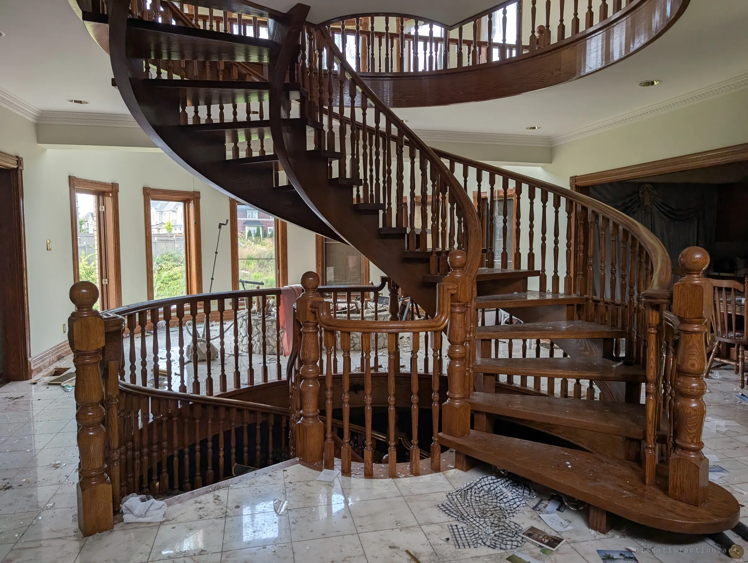 A sweeping open-tread wooden spiral staircase rising through the foyer, with a second curved flight dropping to the basement. Broken glass and family snapshots are scattered across the marble floor.