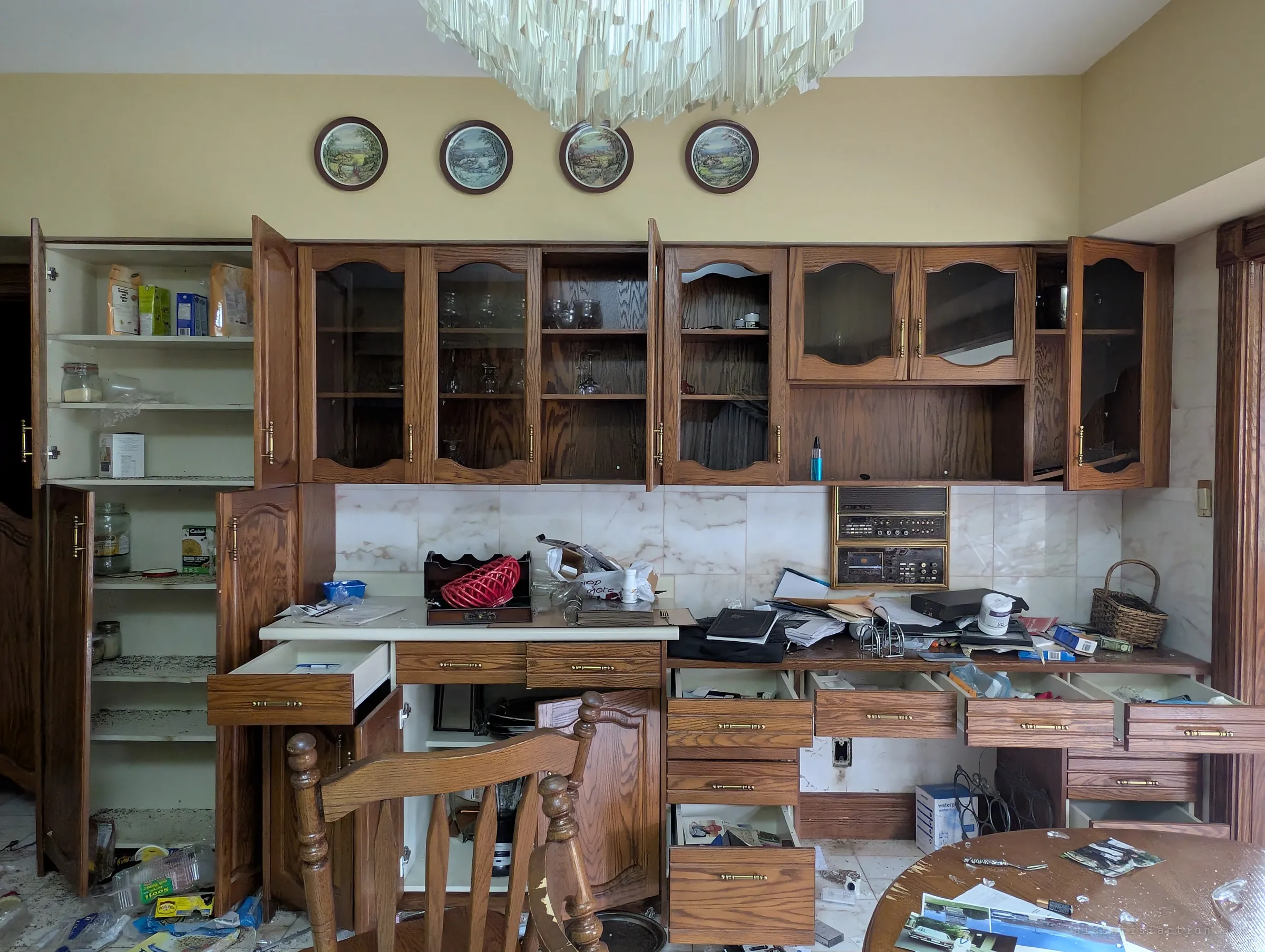 Oak kitchen cabinets hanging open under a row of decorative plates and a crystal chandelier, with drawers pulled out and papers heaped on the counter. A built-in radio and cassette unit still sits in the backsplash.