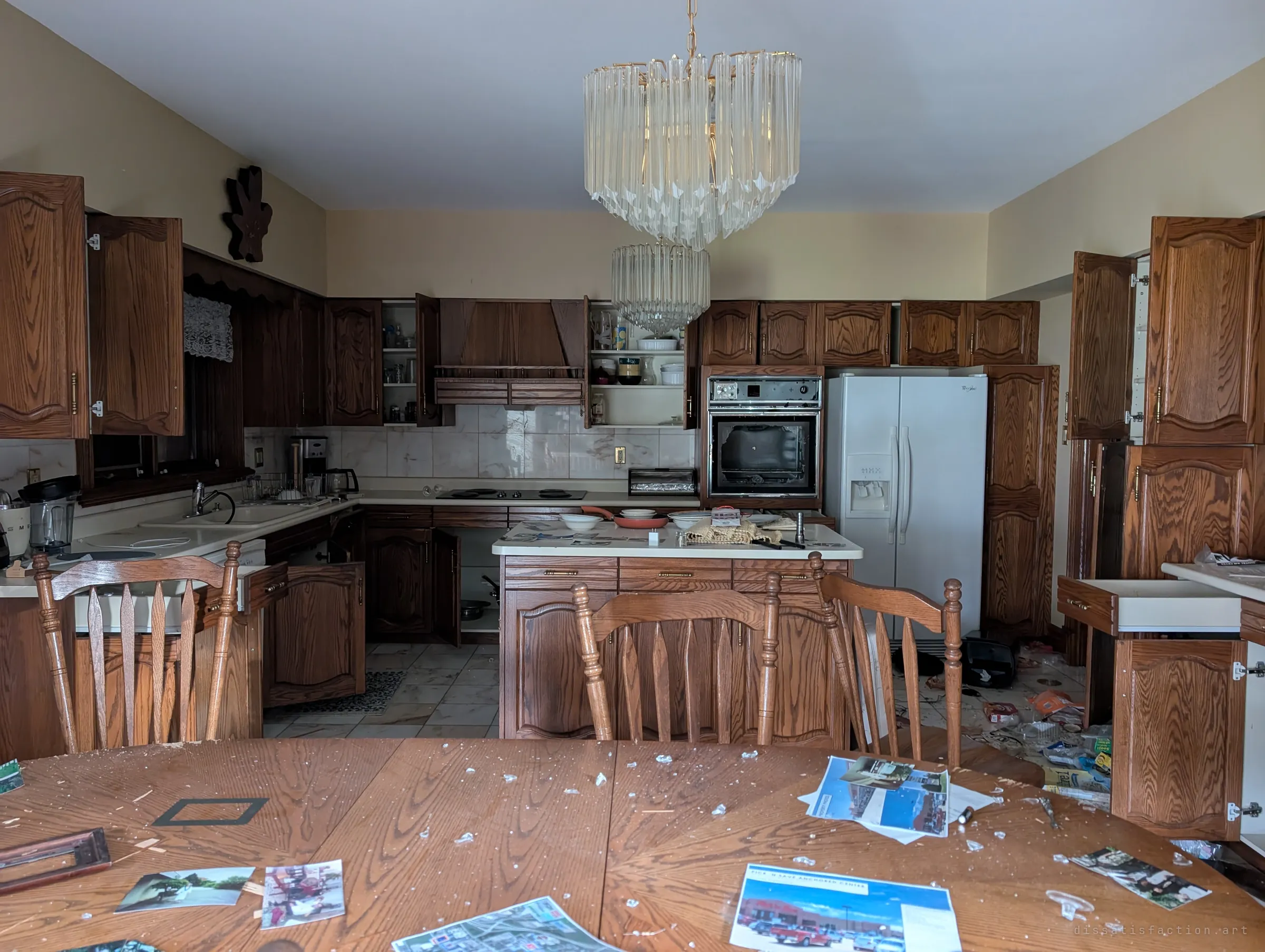 The kitchen seen from the dining table, cabinets thrown open around the island and a broken wall oven, under two tiers of crystal chandeliers. The table is covered in shattered glass and old real-estate printouts.