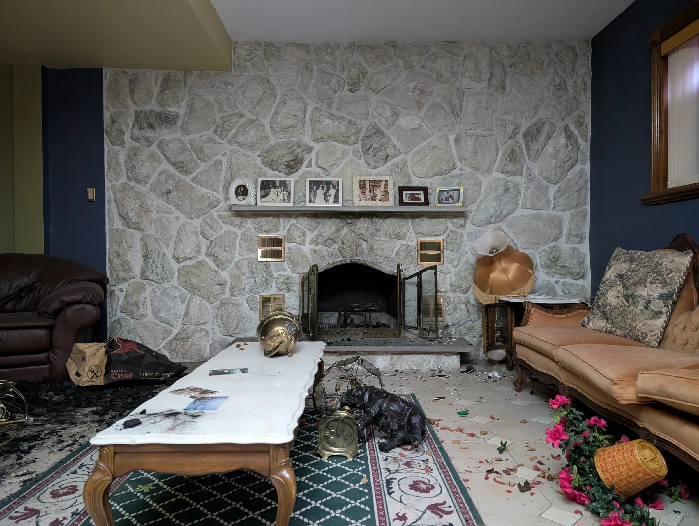 A fieldstone fireplace wall in the basement living room, with framed wedding and family photos still lined up on the mantel. Charcoal, flowers and brass ornaments are scattered across the rug and floor.