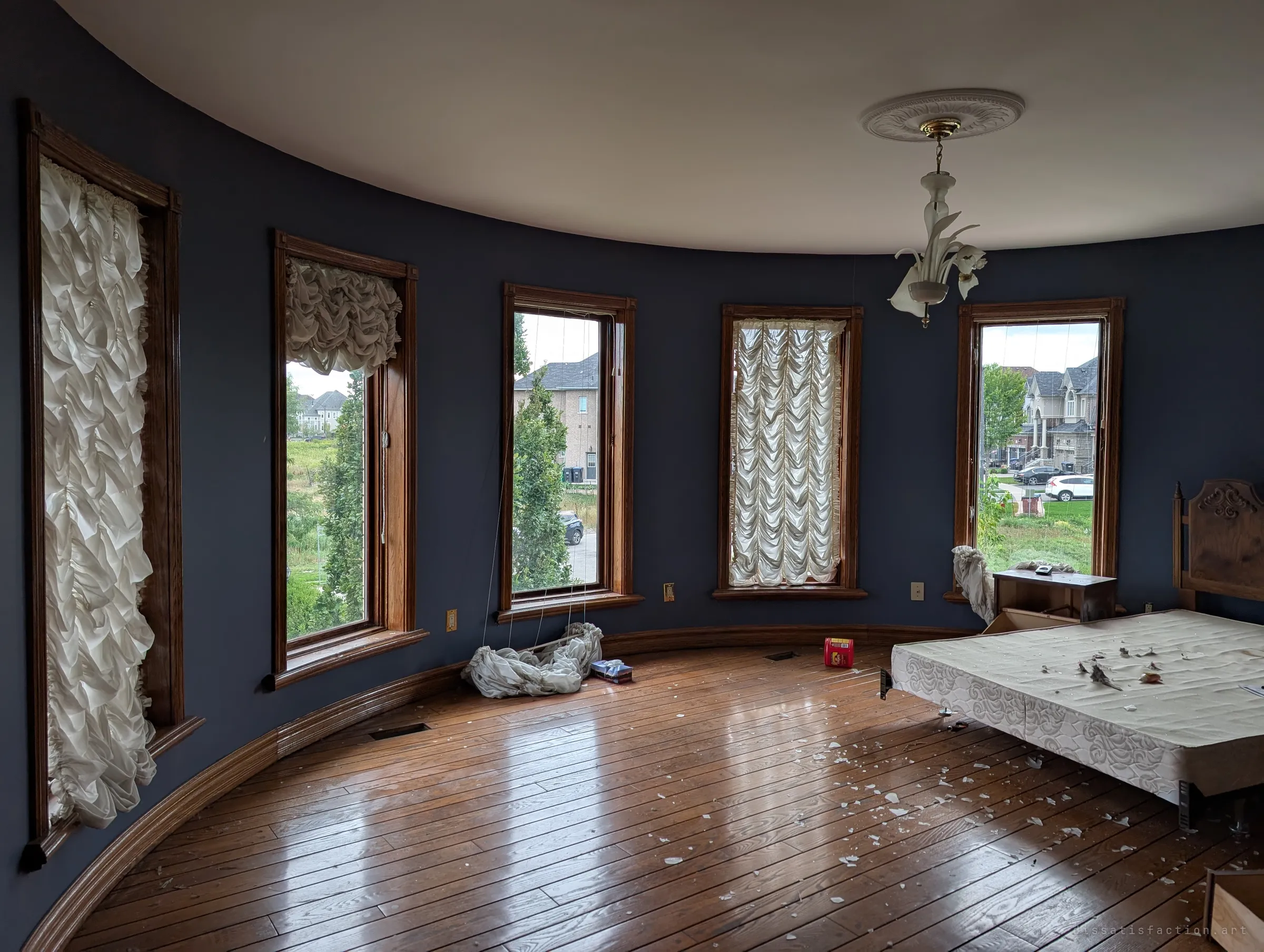 A curved bedroom inside the turret, dark blue walls lined with tall windows and gathered white blinds, a bare mattress base on the floor. Plaster flakes are scattered across the glossy hardwood.