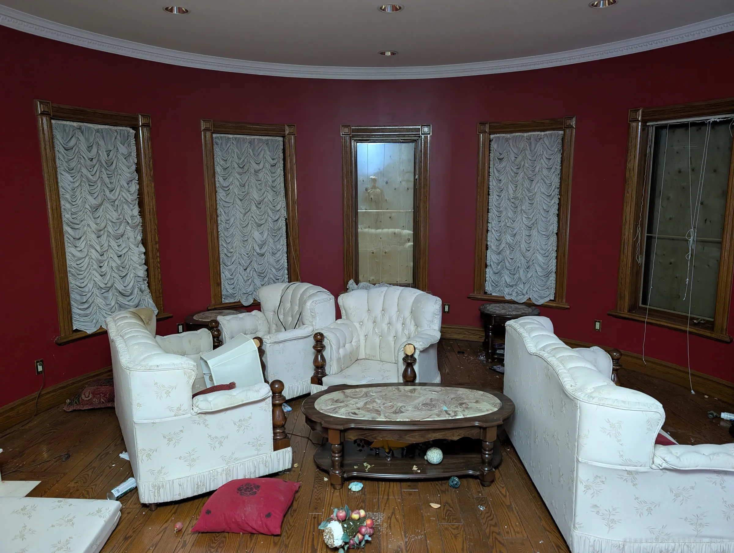 A round red sitting room inside the turret, with cream tufted armchairs around a marble-topped table and ruched blinds over boarded windows. The furniture still stands in place amid the debris.