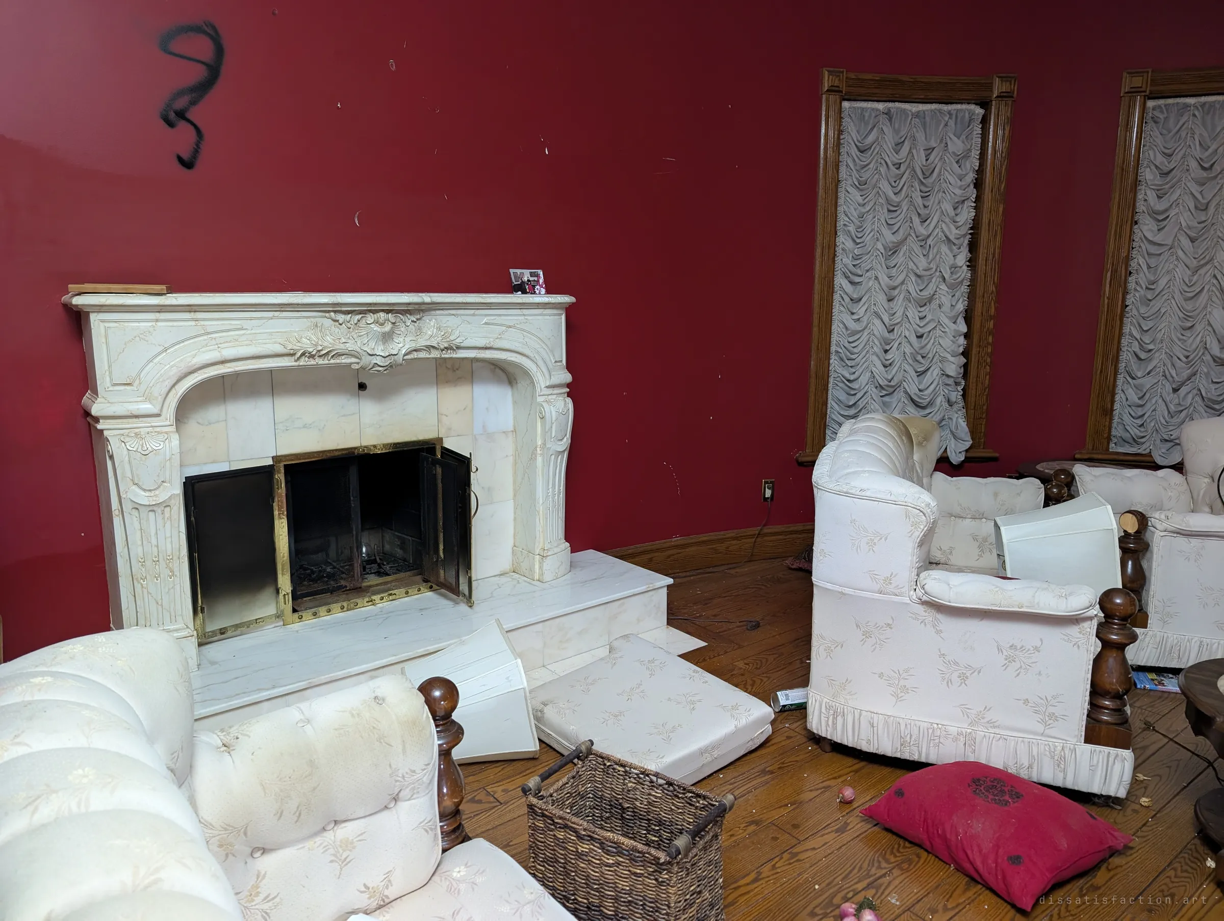 An ornate carved marble fireplace against the red wall of the sitting room, its brass doors hanging open. Lampshades and cushions have been knocked to the floor, and a single graffiti squiggle marks the wall.