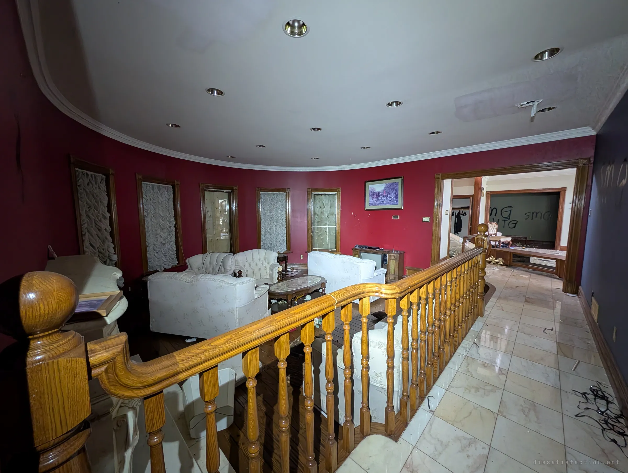 Looking down over a wooden banister into the sunken red sitting room, with a hallway of marble tiles leading off to a graffiti-covered room. A hunting print and an old console TV are still against the walls.