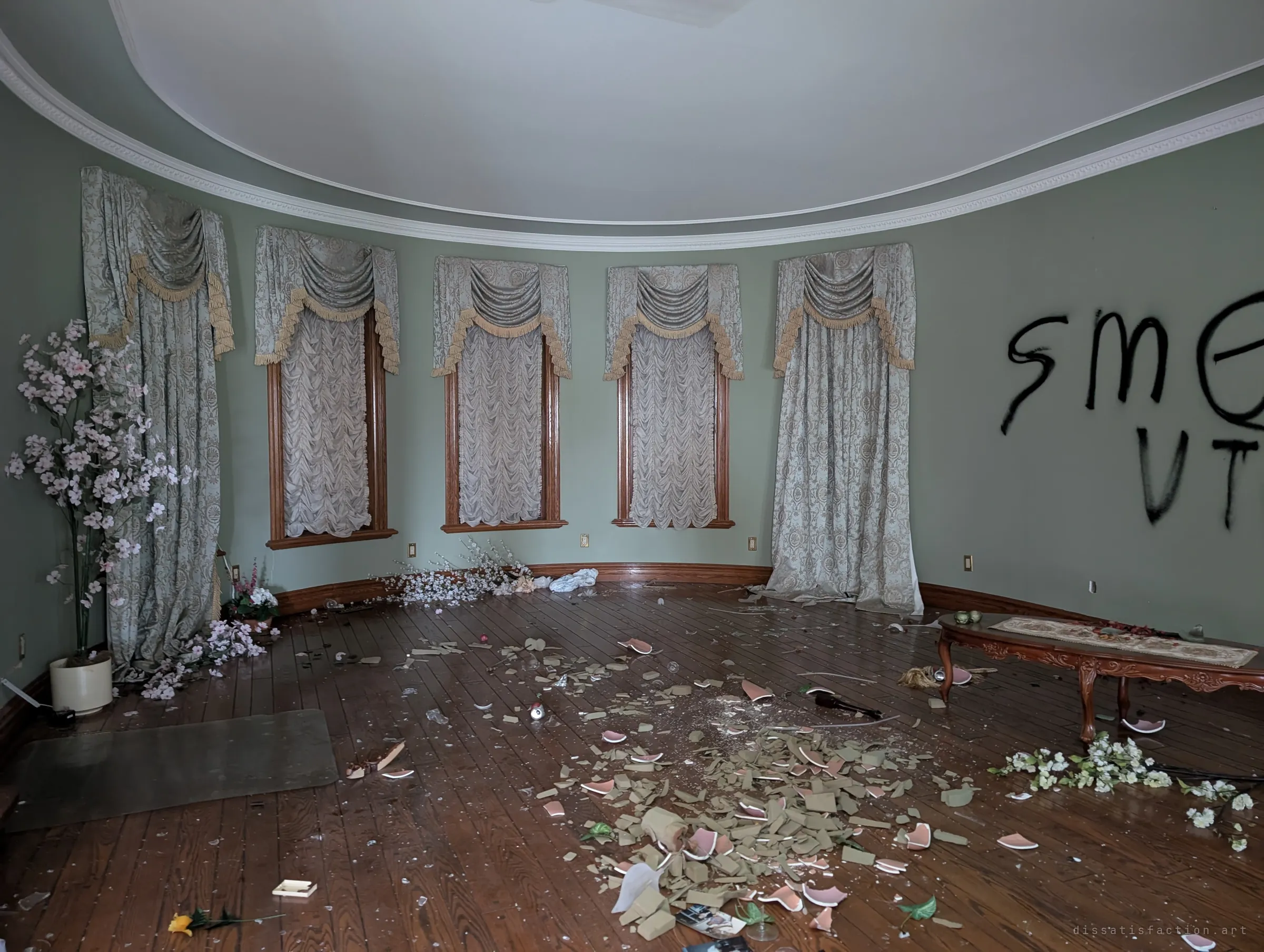 A curved drawing room with draped valance curtains over tall windows, spray-painted graffiti on the green wall and shattered pottery and silk flowers across the hardwood floor. The ornate finishes make the vandalism feel all the more abrupt.