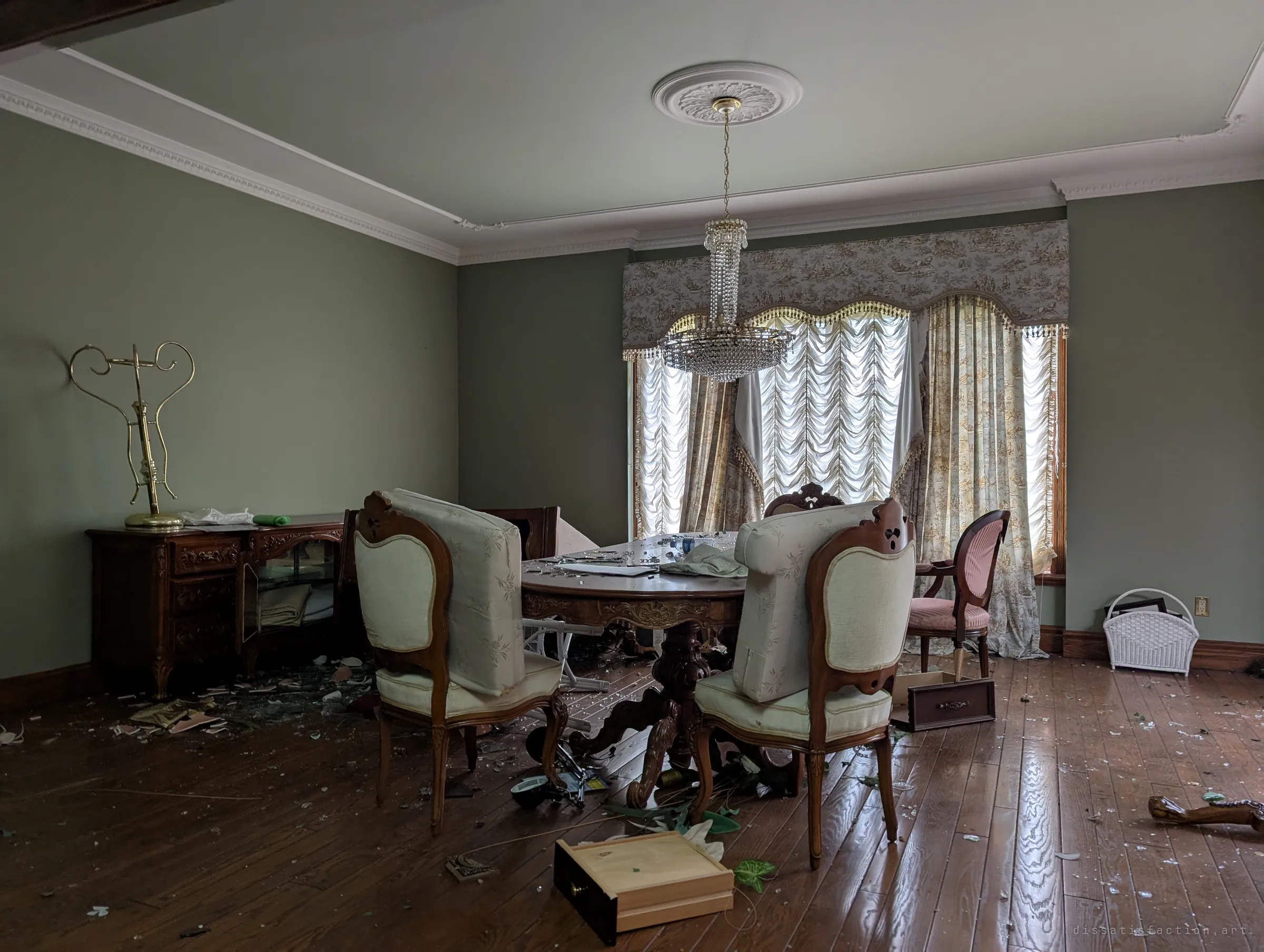 A formal dining room with a crystal chandelier over a carved pedestal table, upholstered chairs and a pulled-out drawer lying in broken glass. Daylight filters through ruched curtains onto the abandoned setting.