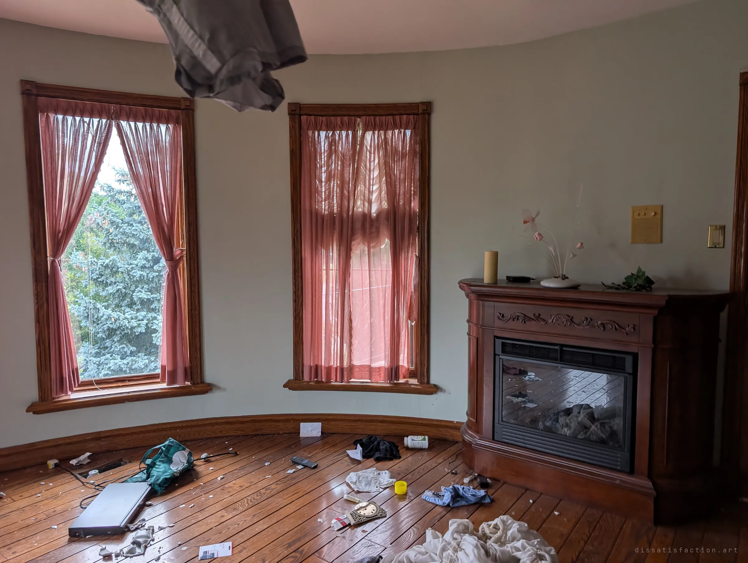 A rounded bedroom corner with pink sheer curtains, a wooden electric fireplace and household items scattered across the floor. A bedsheet hangs down from the ceiling in the foreground.