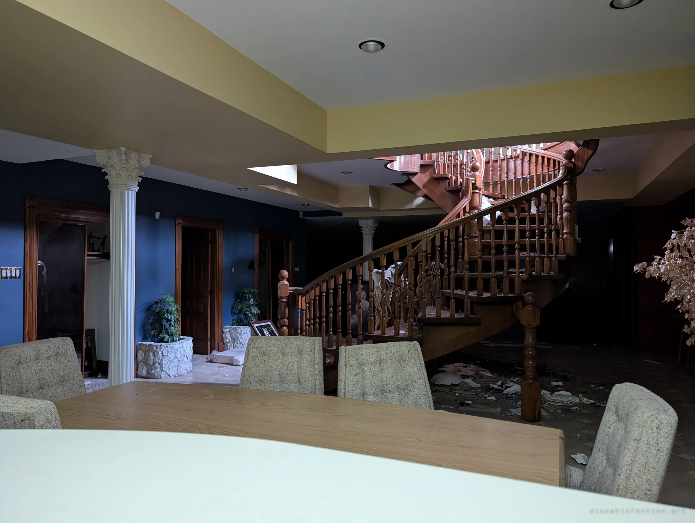 A lower-level lounge seen past a table and upholstered chairs, with a carved wooden spiral staircase curving up beside white Corinthian columns. Debris gathers beneath the stairs in the dim light.
