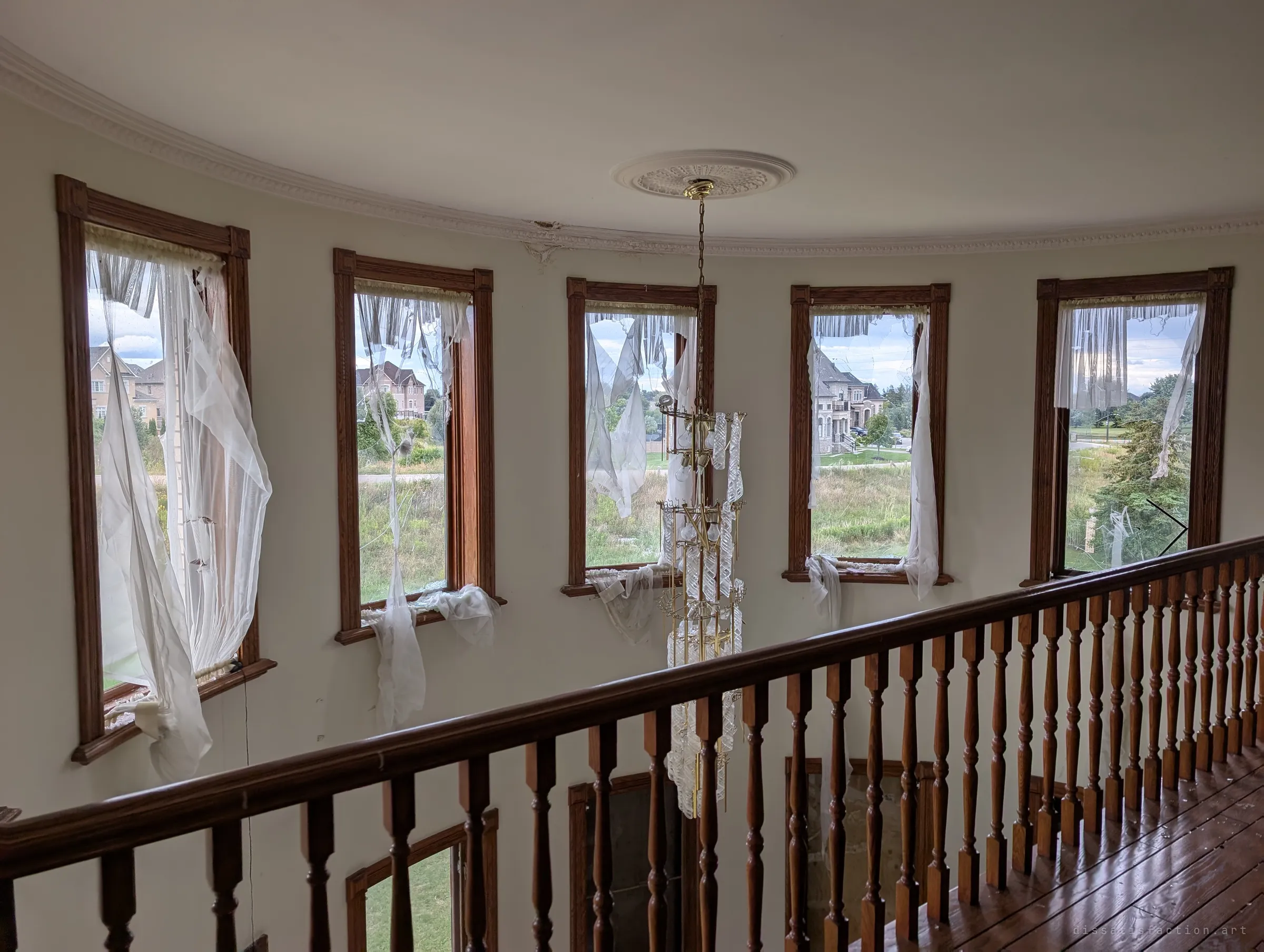 From an upper gallery railing, a tall rounded wall of windows with torn sheer curtains and a long crystal chandelier dropping into the stairwell. Light floods in from outside.
