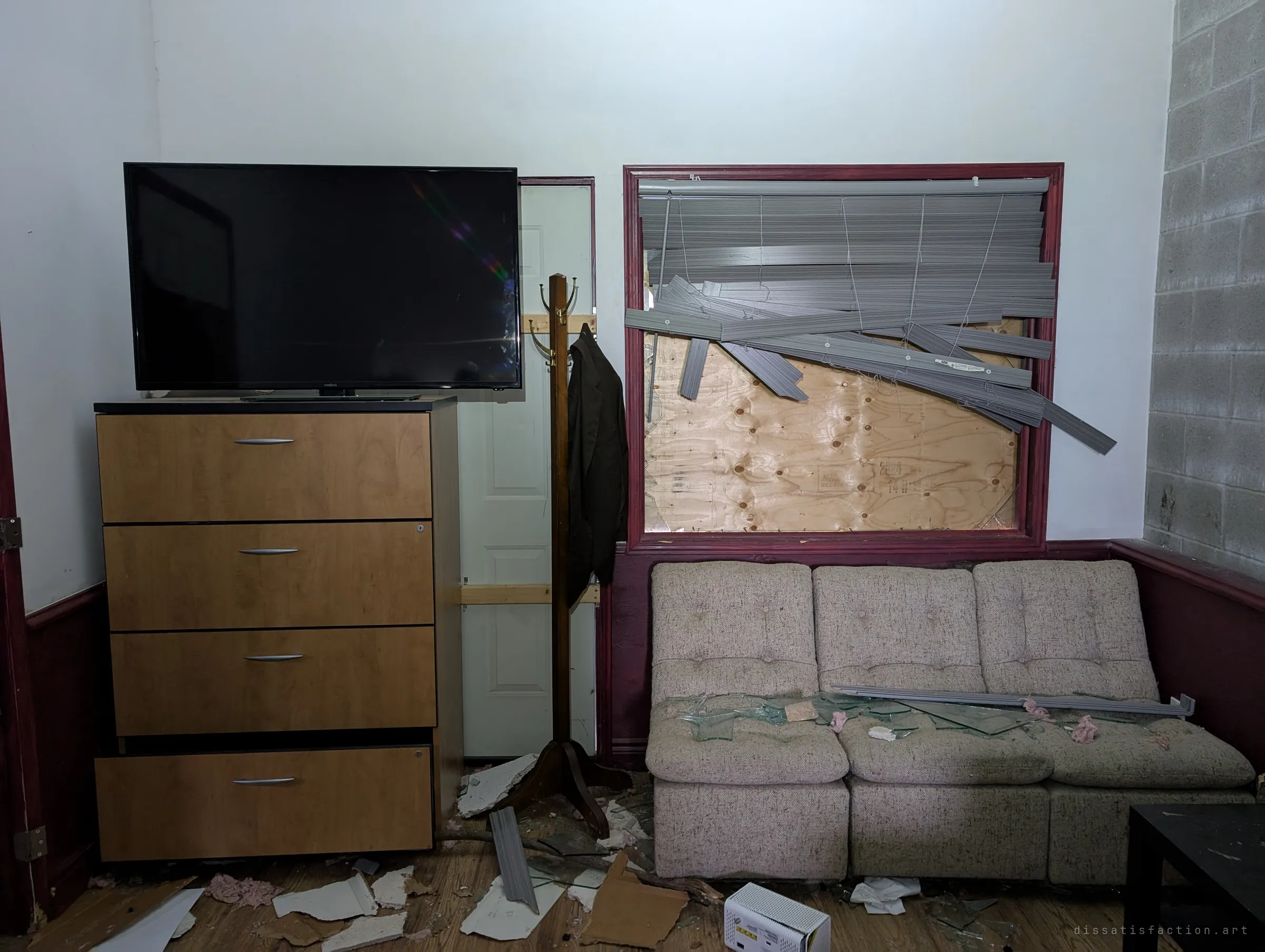 A small room with a flat-screen TV on a filing cabinet, a coat stand with a jacket still hanging, and a boarded window whose blinds were torn down onto a speckled sofa. Broken glass and ceiling pieces litter the floor.
