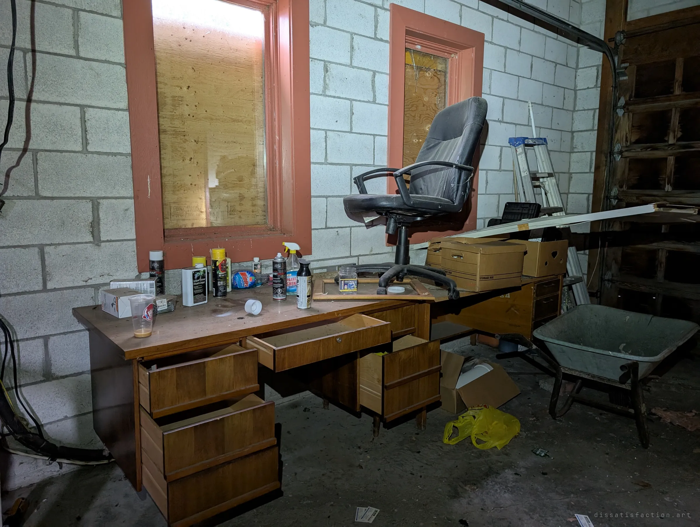 An old wooden desk against a cinder-block wall with its drawers pulled open, spray cans and cleaners on top and an office chair perched on a second desk. Boarded windows and a wheelbarrow complete the workshop scene.